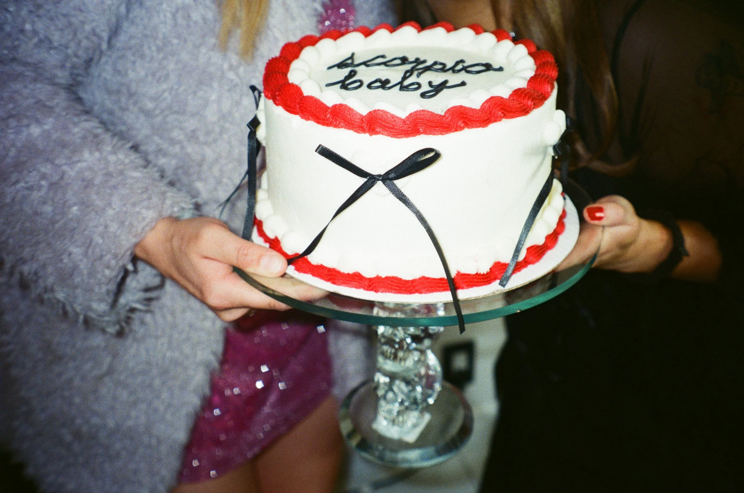 A birthday cake with white icing, decorated with a red border and a black ribbon design, being held by two people. The cake has a message written on top in dark frosting, but the writing is unclear.