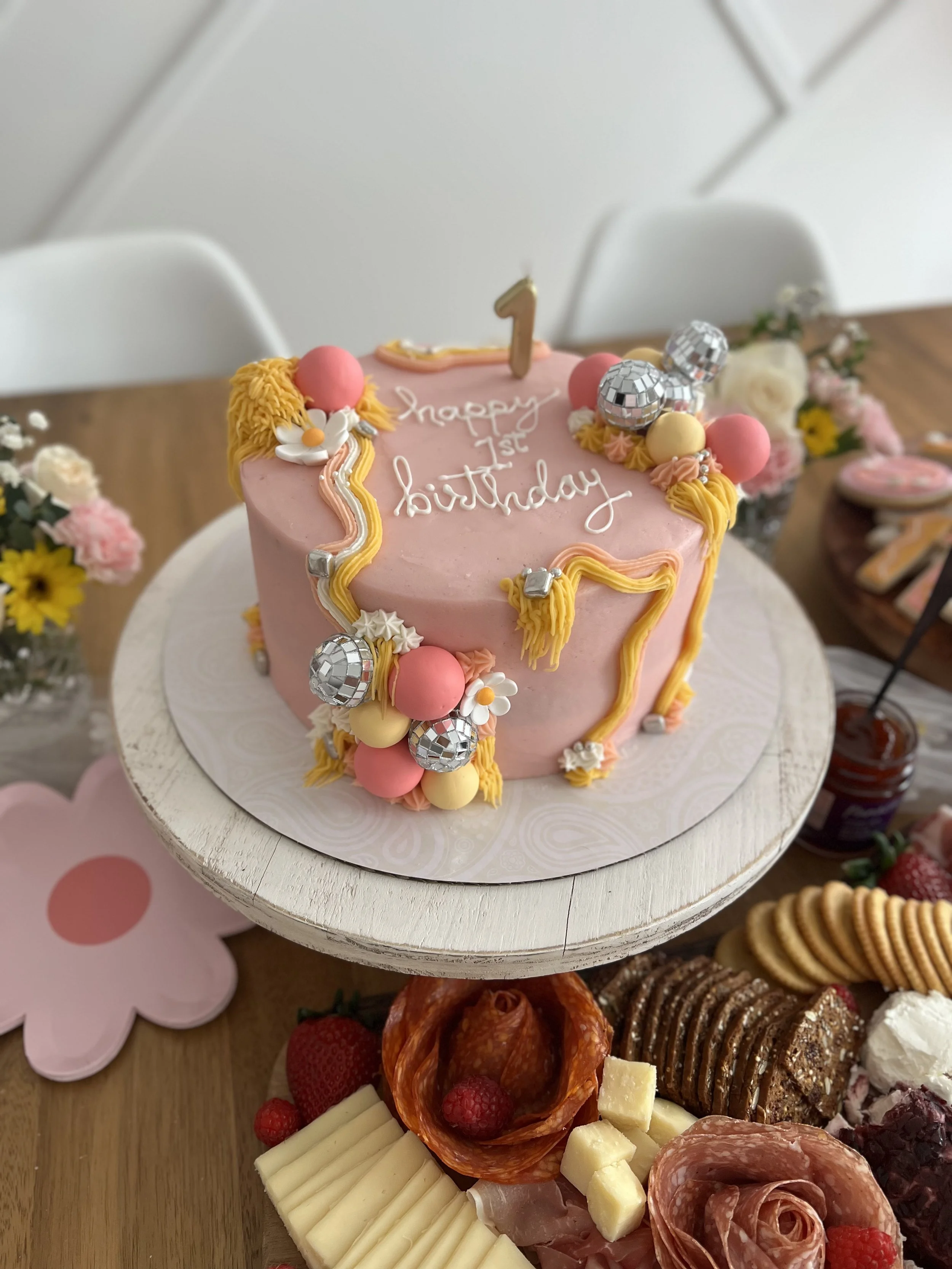 Pink birthday cake with a gold number 1 topper and white writing that says 'happy 1st birthday', decorated with yellow, pink, and silver accents, placed on a white stand. Surrounding the cake are various cheeses, crackers, cured meats, strawberries, 
