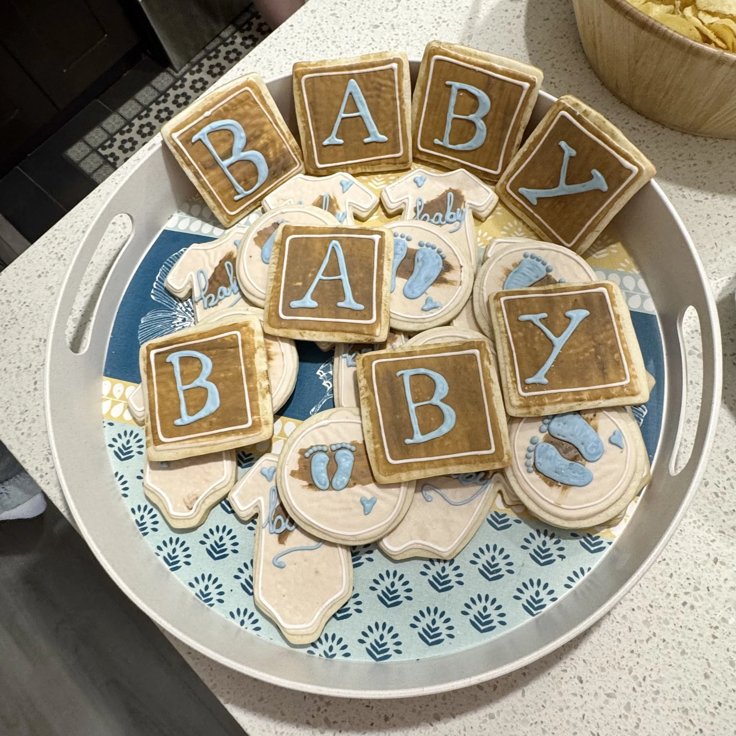 Decorated cookies with blue letters forming the word 'BABY' placed on a tray with a blue and white patterned paper, on a light-colored kitchen countertop.
