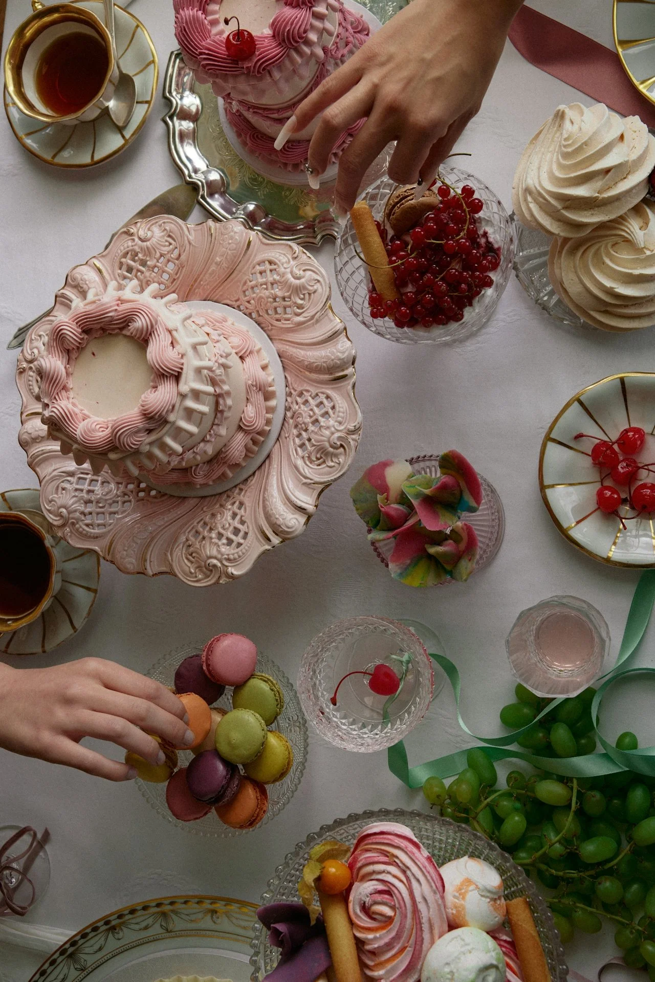 A table with various desserts including pink and white frosted cakes, macarons, meringues, and cherries, with a person's hand reaching for colorful macarons.