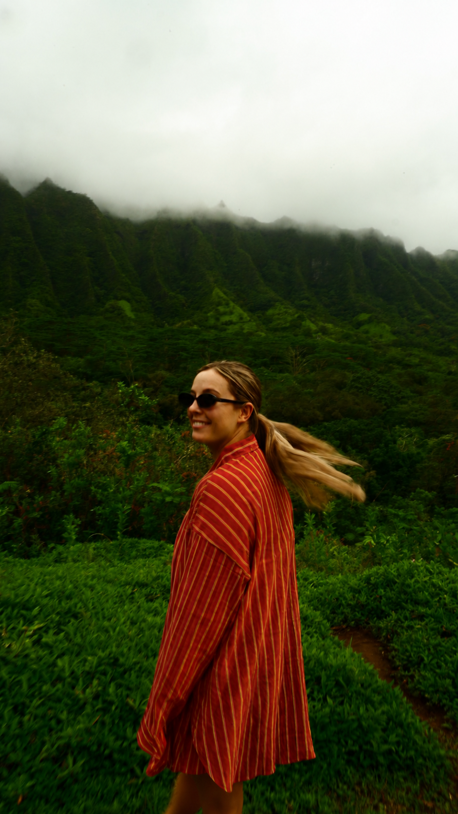 A woman with blonde hair wearing sunglasses and a red, yellow, and brown striped shawl is smiling in front of lush green mountains shrouded in clouds.