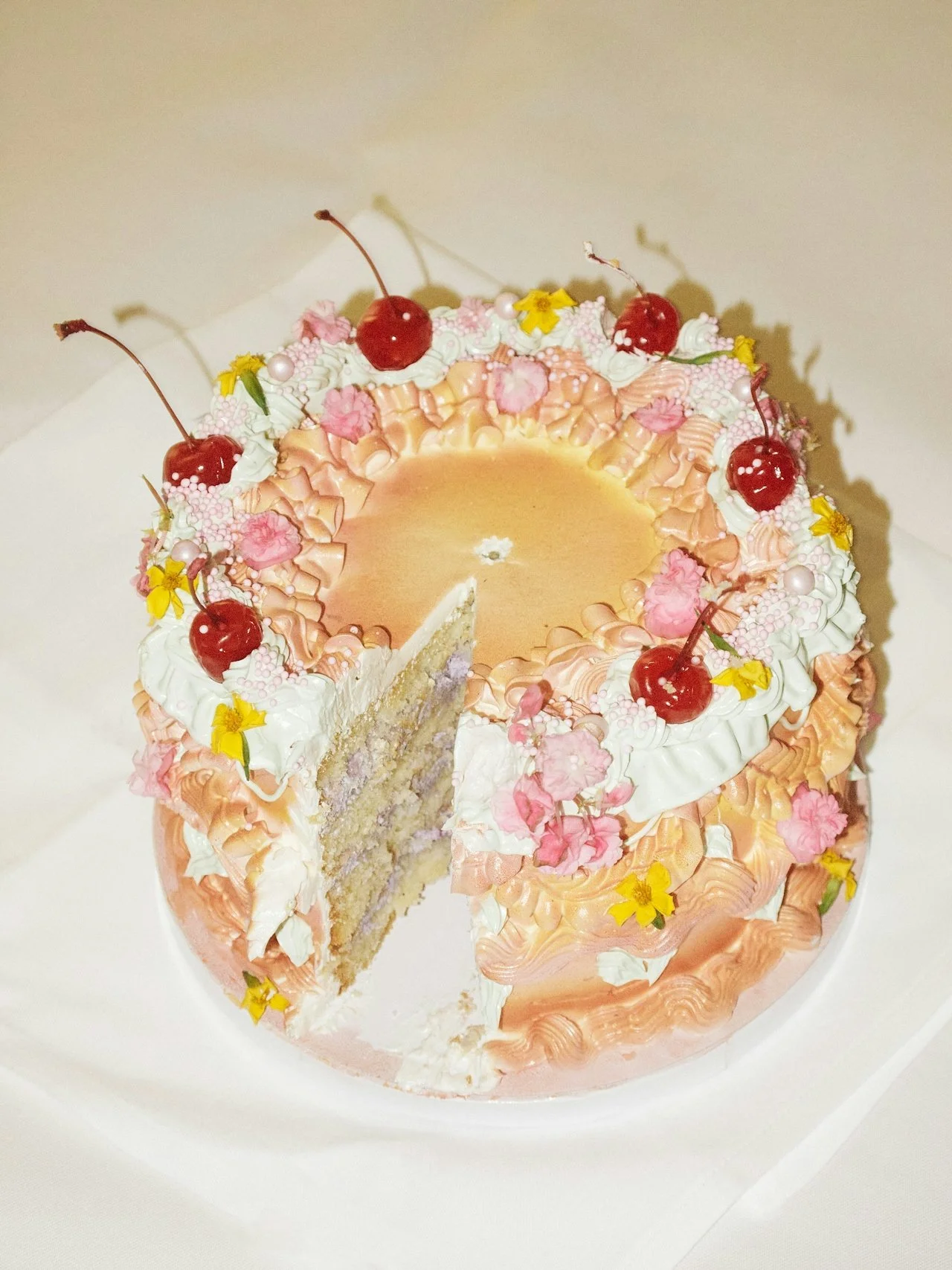 A round cake decorated with pink and white icing, cherries on top, yellow flowers, and pink decorations, with a slice removed showing the inside layers.