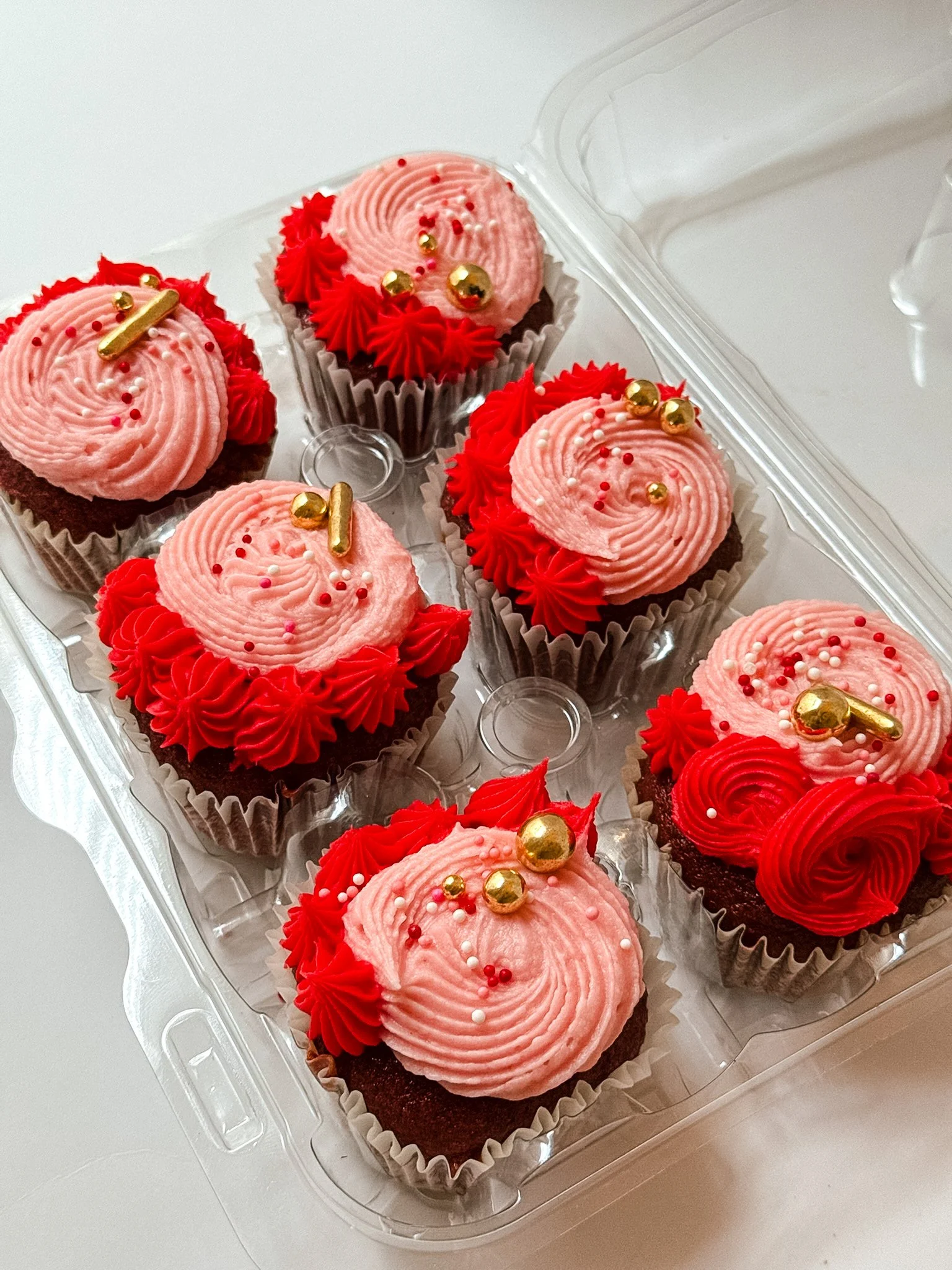 A clear plastic container holding six decorated cupcakes with pink and red frosting, gold and red spherical sprinkles, and some with gold candy bars on top.