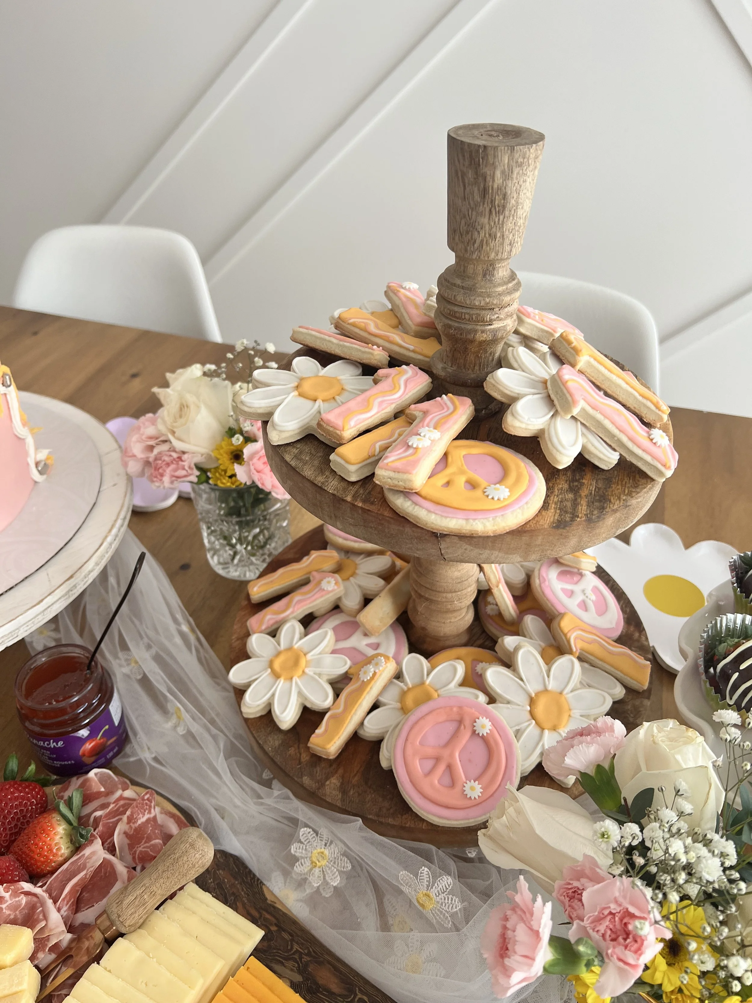 Decorative cookies with floral designs on a wooden cake stand, surrounded by flowers, cheese, and charcuterie on a table.