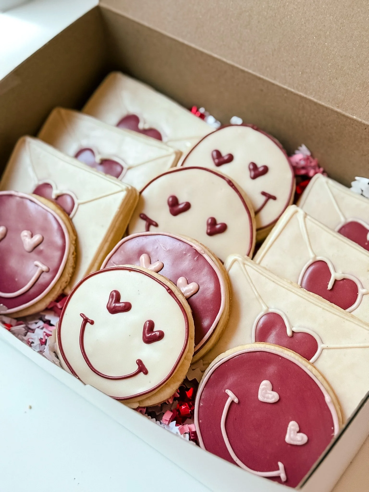 A box of decorated cookies with heart and smiley face designs, some with pink and white icing.