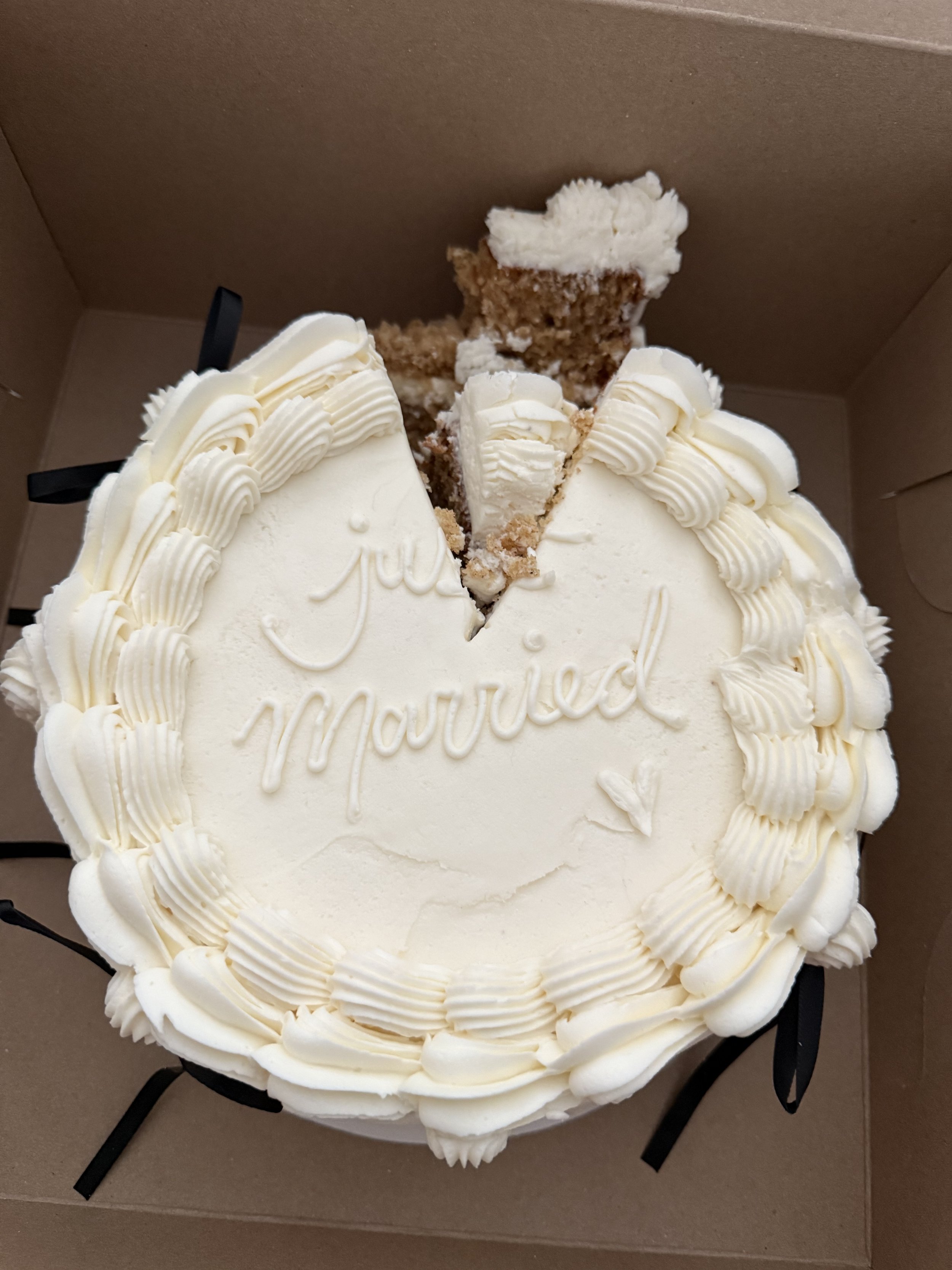A round white cake with cream piping around the edges and a slice cut out, showing brown and white interior layers, decorated with 'Happy Mother's Day' written in icing.