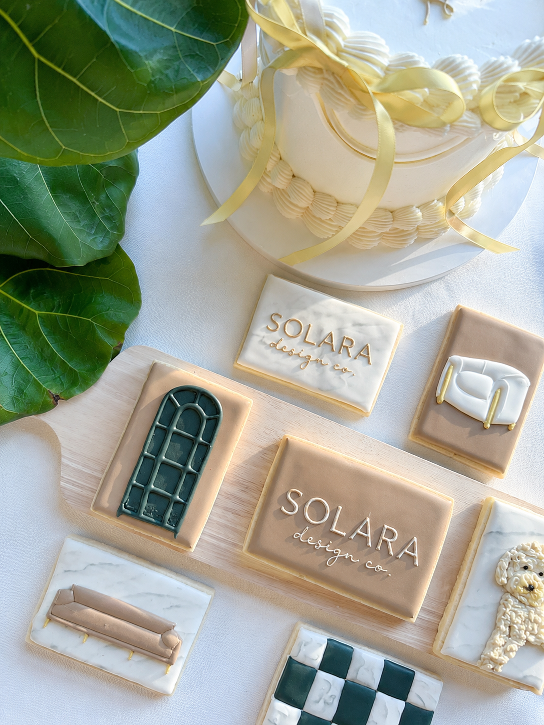 Decorative cookies with designs and text, a white frosted cake with yellow ribbon details, green leaves, and a white card with gold lettering that reads 'SOLARA design co.'.