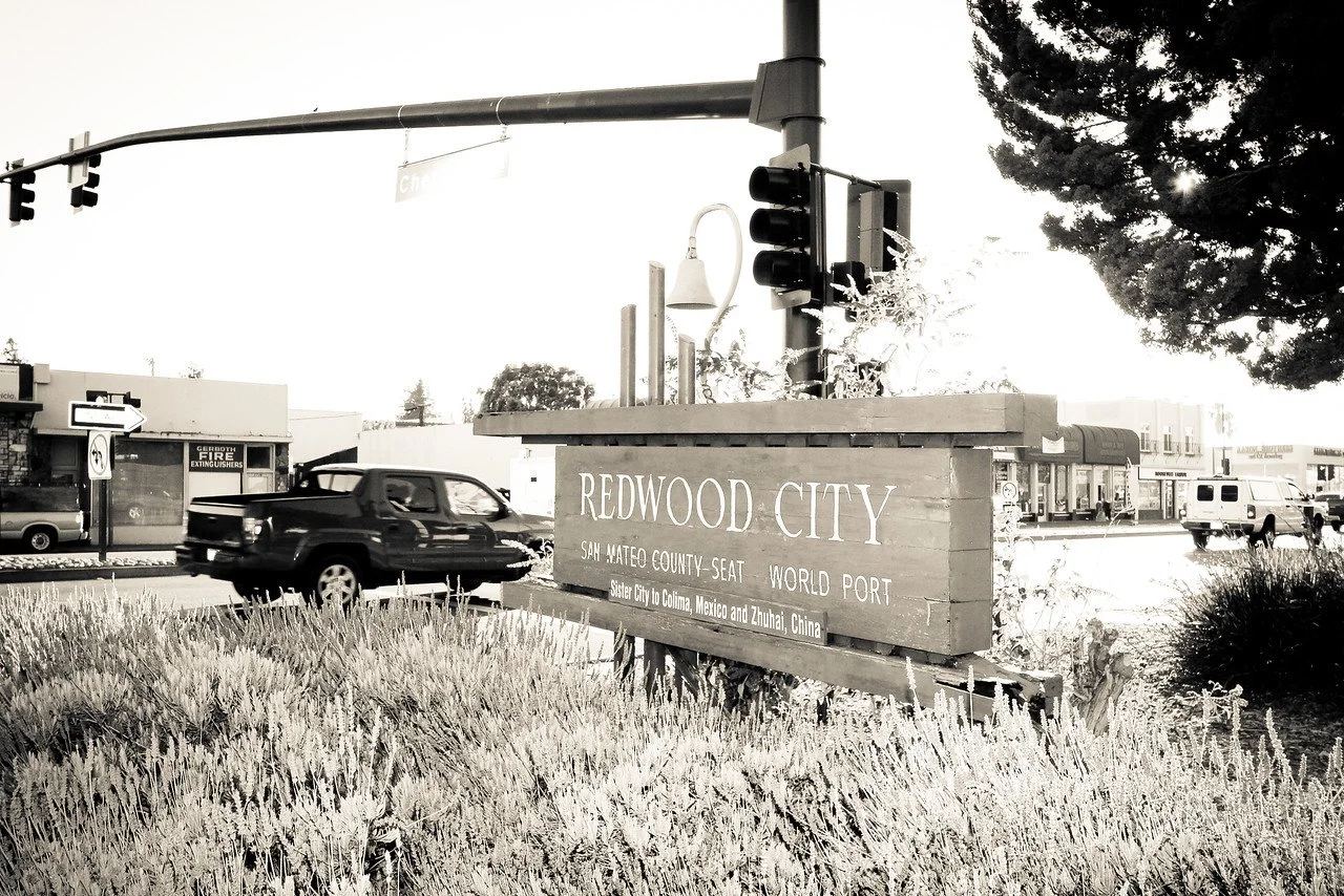 Black and white photo of a street corner with a sign that reads "Redwood City" and mentions sister cities in Mexico and China, with cars passing by and traffic lights overhead.