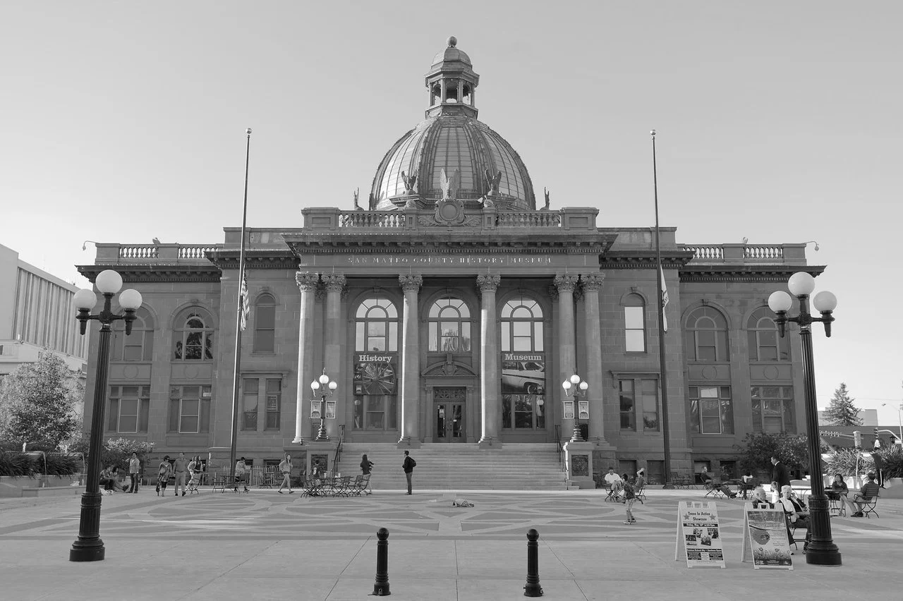 Black and white photo of a historic government building with a large central dome, columns at the entrance, and people walking and sitting outside.