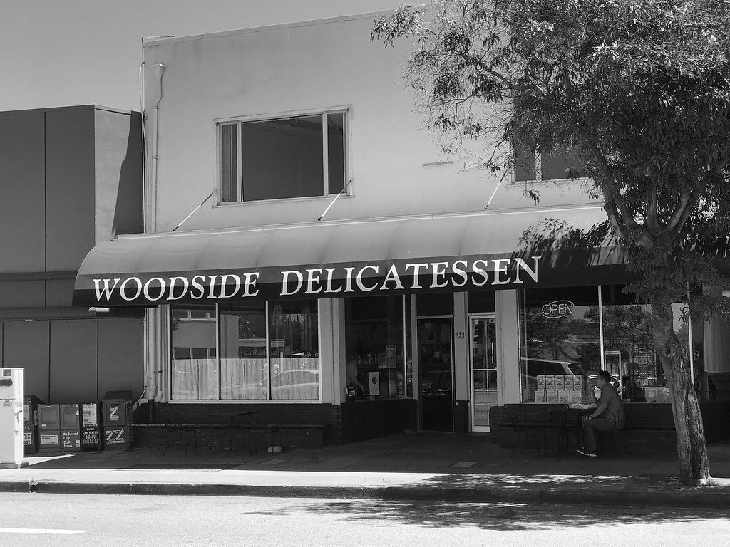 A storefront with a sign that reads 'WOODSIDE DELICATESSEN' in bold letters. There are benches outside, one person sitting at a bench, a small tree on the sidewalk, and the store has an 'OPEN' sign in the window.