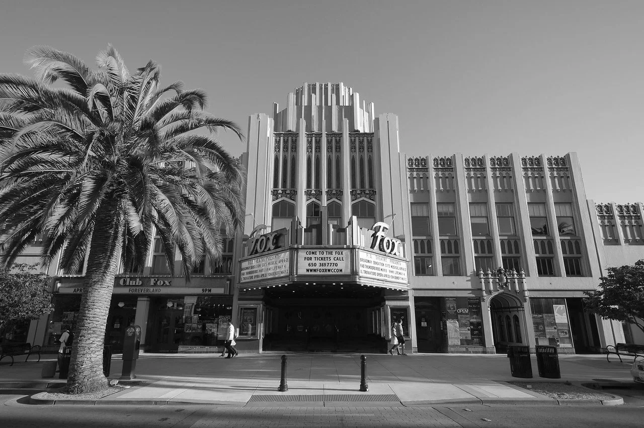 Black and white photo of a theater building with a marquee sign, palm tree in the foreground, and pedestrians on the sidewalk.