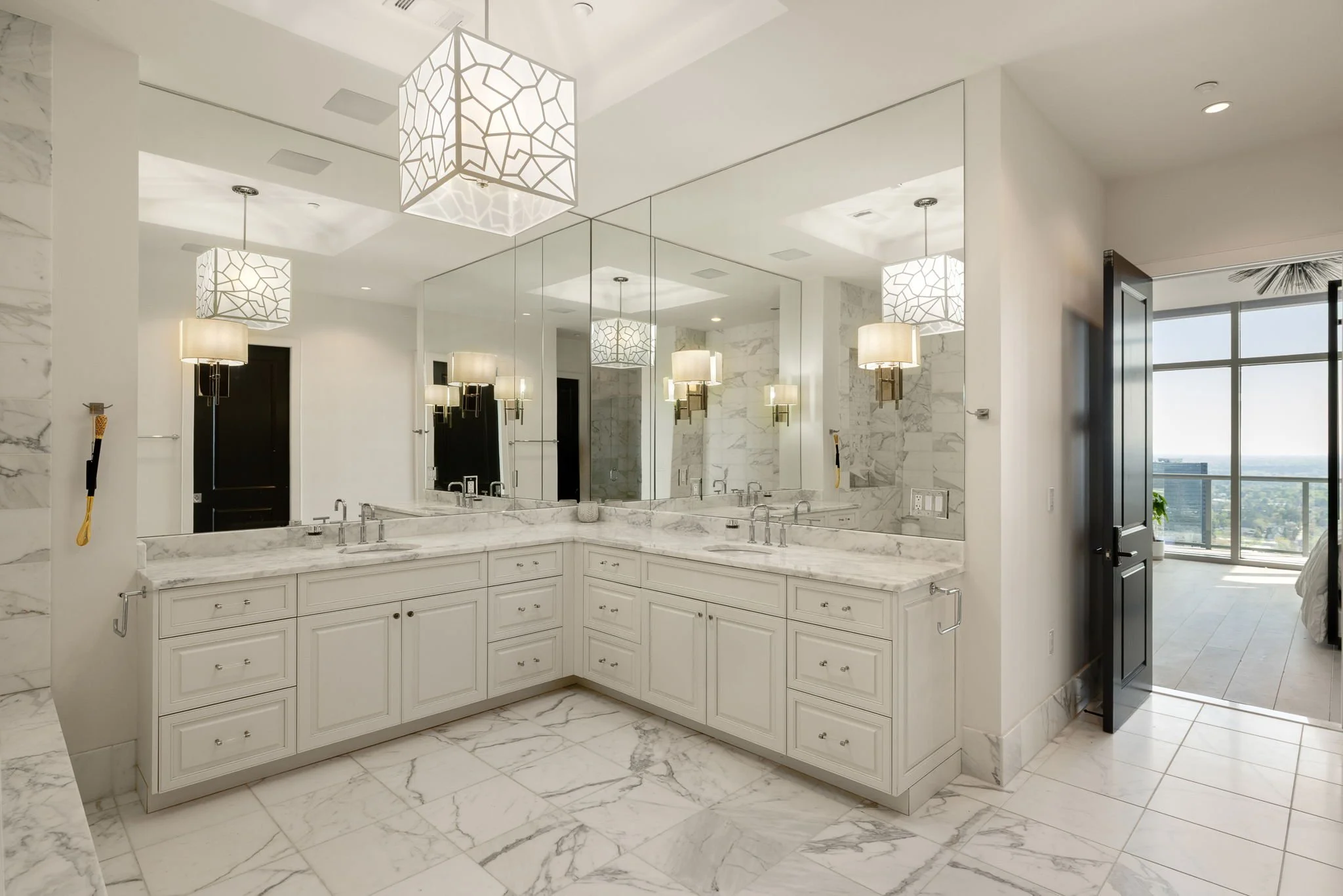 Modern bathroom with white marble countertops, multiple sinks, large mirror, wall-mounted light fixtures, and an open door leading to a room with a balcony and city view.