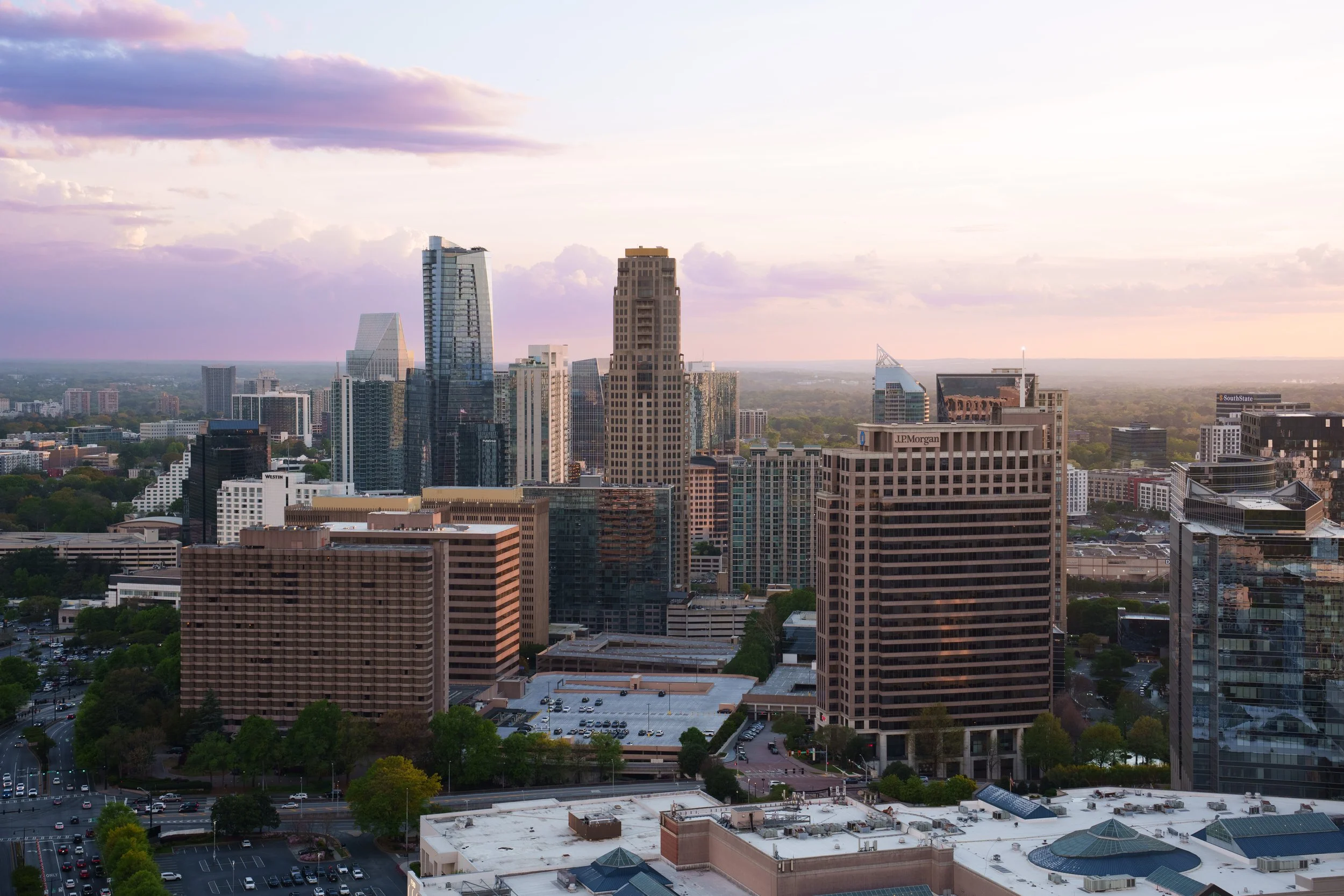 A city skyline at sunset with tall skyscrapers, some with glass facades reflecting the sky, and a few lower buildings with parking lots and trees in the foreground.