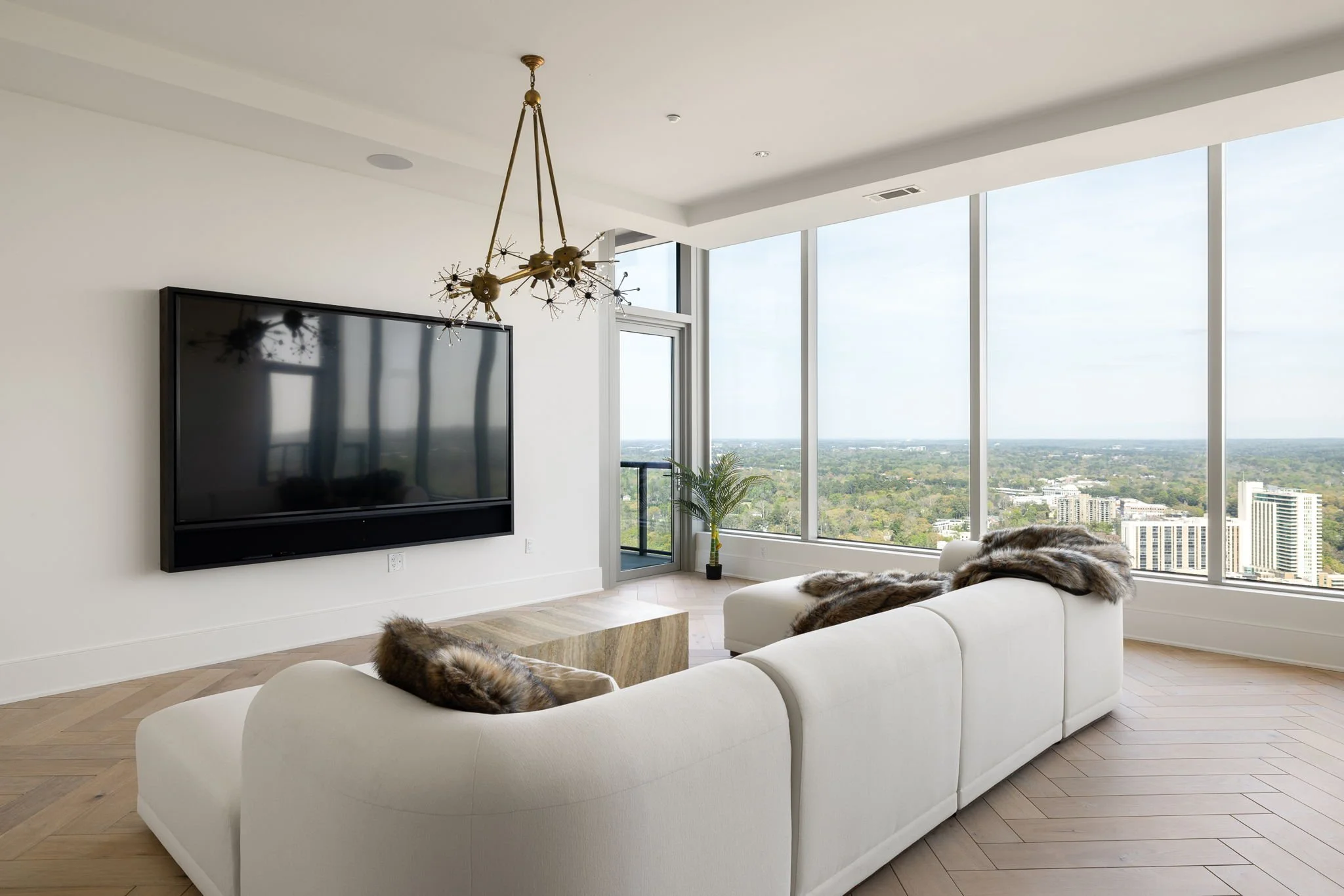 Living room with white sofa, fur throw pillows, large windows showing city view, wall-mounted flat-screen TV, modern chandelier, hardwood flooring, and a potted plant.