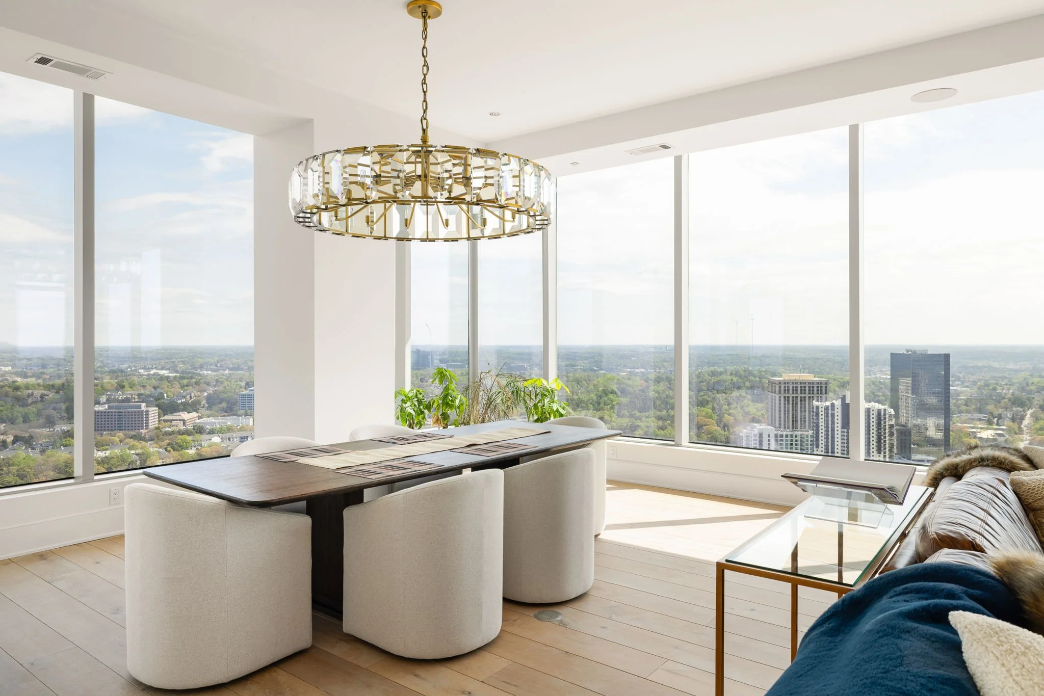 Modern high-rise living room with large windows, a wooden dining table with white upholstered chairs, a sofa with a throw blanket, a glass coffee table, and a chandelier, overlooking a cityscape and greenery.