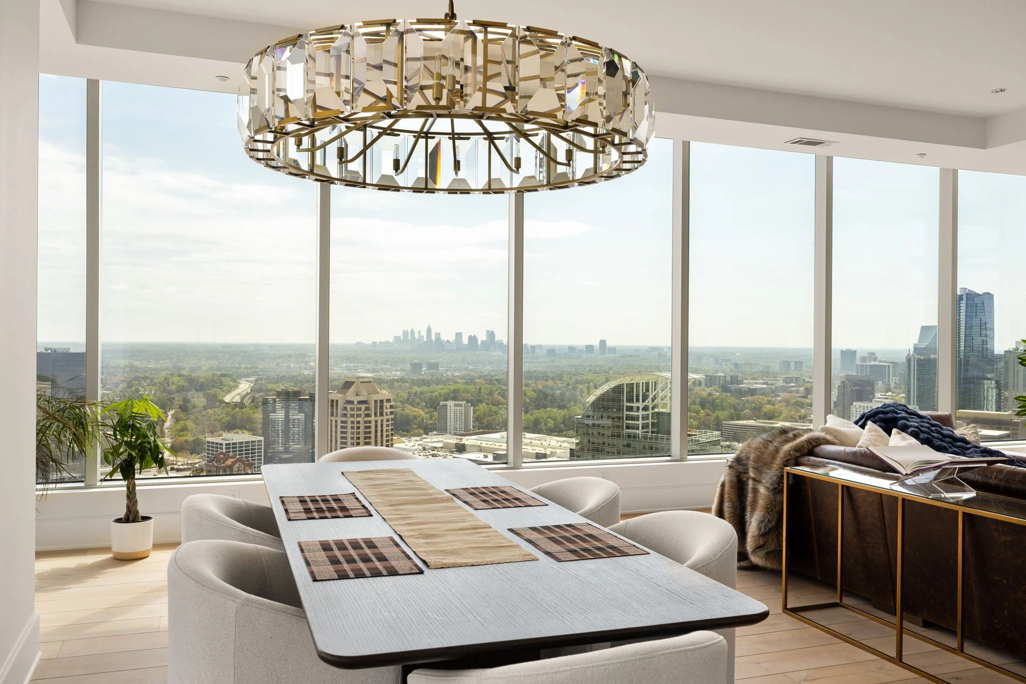 Modern dining room with a white table, four chairs, and a city view through floor-to-ceiling windows, featuring a large crystal chandelier.