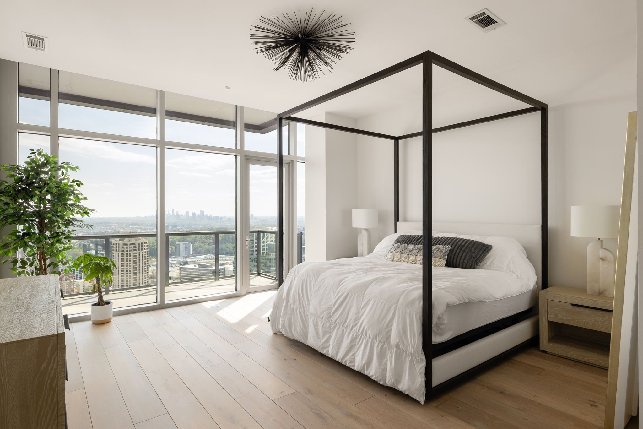Modern bedroom with a large glass window showing city skyline view, white canopy bed with black frame, white bedding, and side tables with lamps.