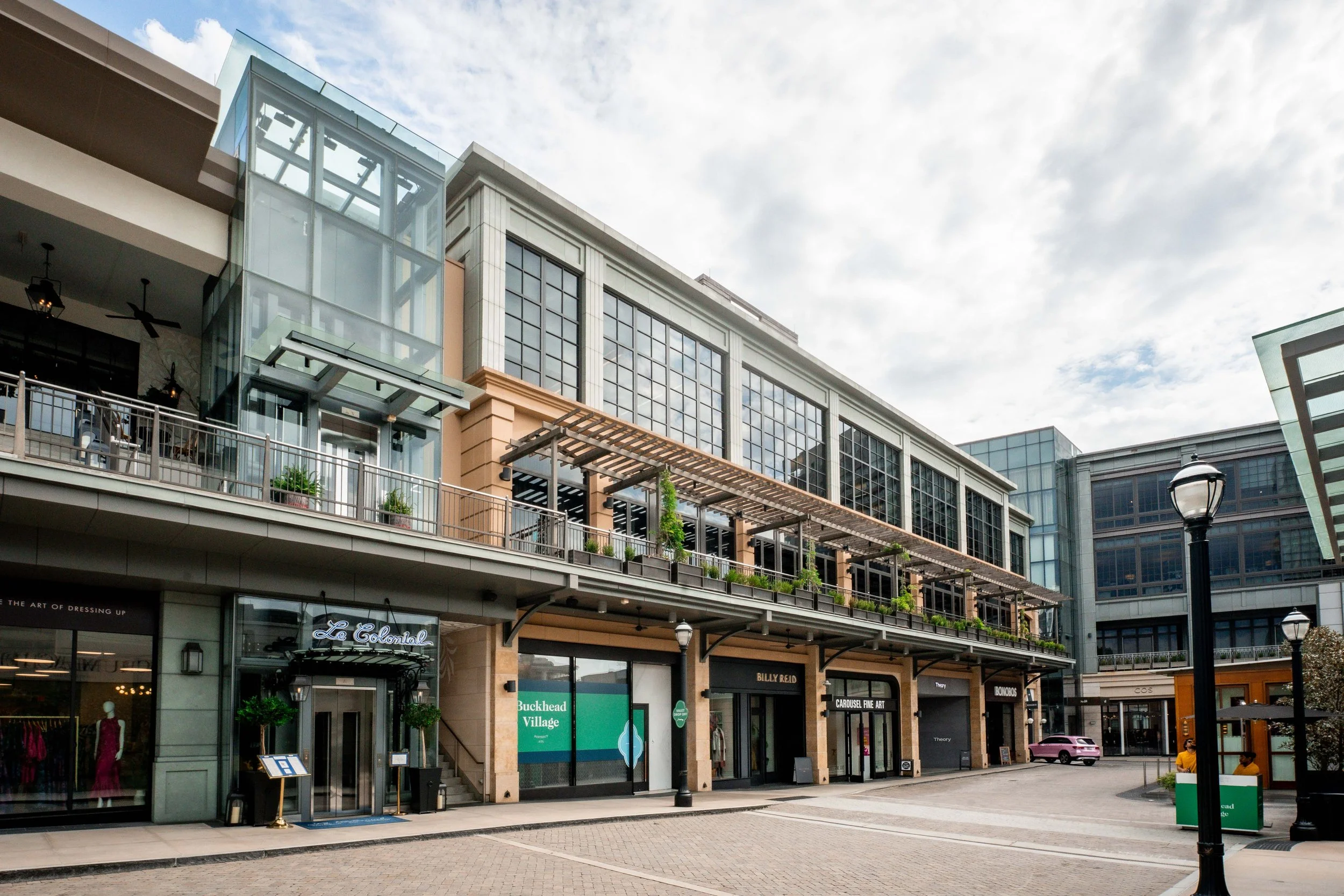 A multi-story shopping plaza with stores, large windows, and outdoor seating areas, under a cloudy sky.