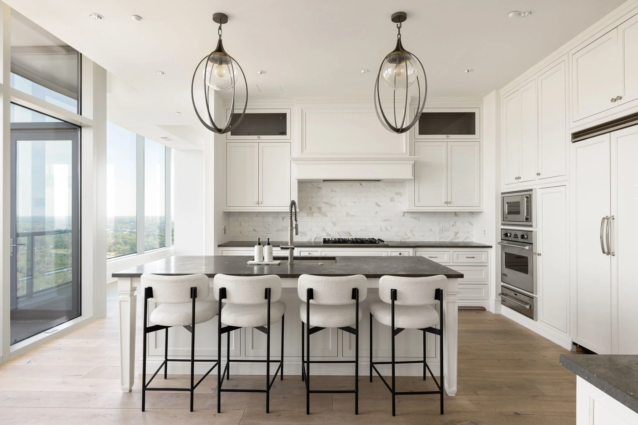 Modern white kitchen with an island, four bar stools, pendant lights, and large windows overlooking a cityscape.