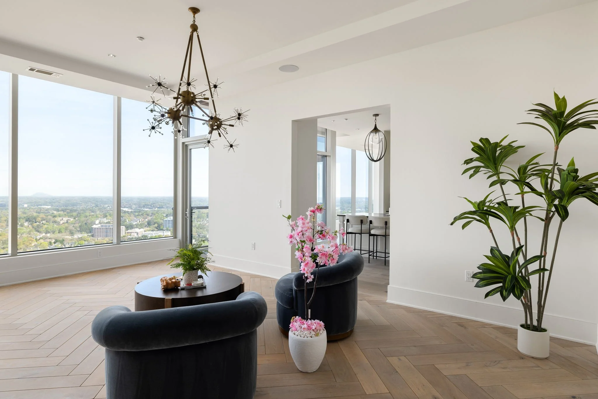 Modern living room with large windows, a chandelier, two dark blue armchairs, a round coffee table with plants, pink flowers, and a large potted plant, overlooking a cityscape.