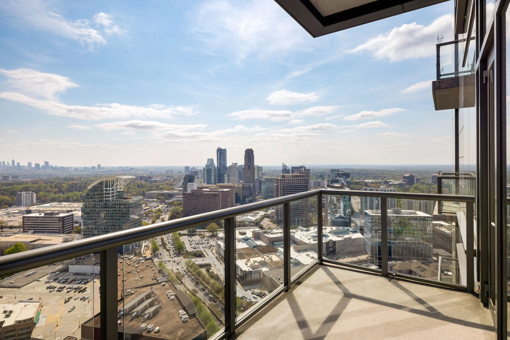 View from a high-rise balcony overlooking a city skyline with tall buildings, streets, and parking lots under a partly cloudy sky.
