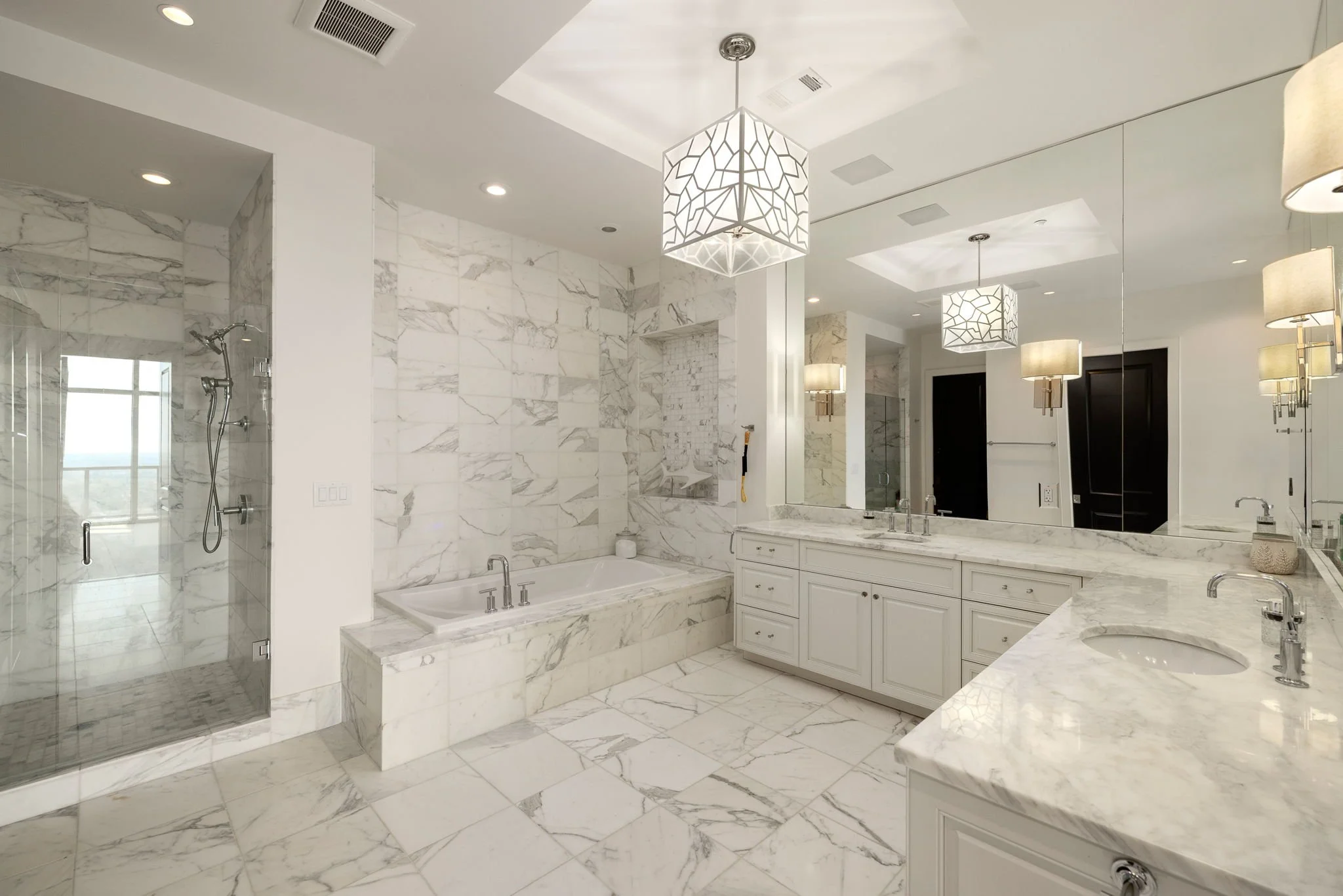 Spacious luxury bathroom with marble tiles, bathtub, glass-enclosed shower, double vanity with marble countertop, large mirror, pendant and wall sconce lighting, with window reflection in mirror.