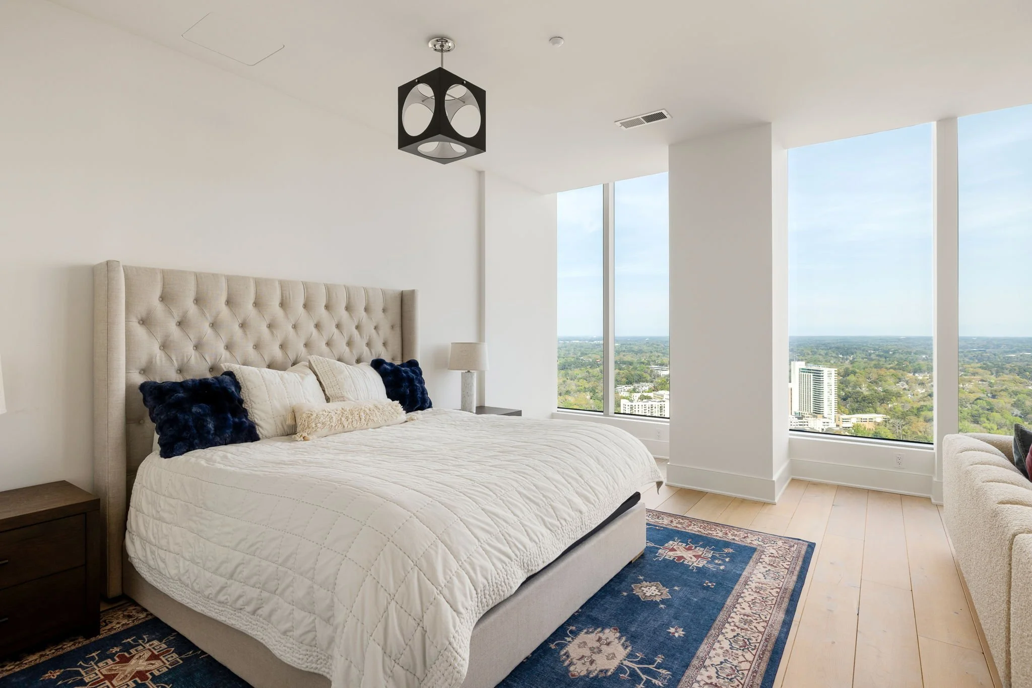 Modern bedroom with large windows offering city and green views, beige tufted headboard, cream bedding, blue and beige pillows, wooden nightstands, a small lamp, and a blue patterned area rug.