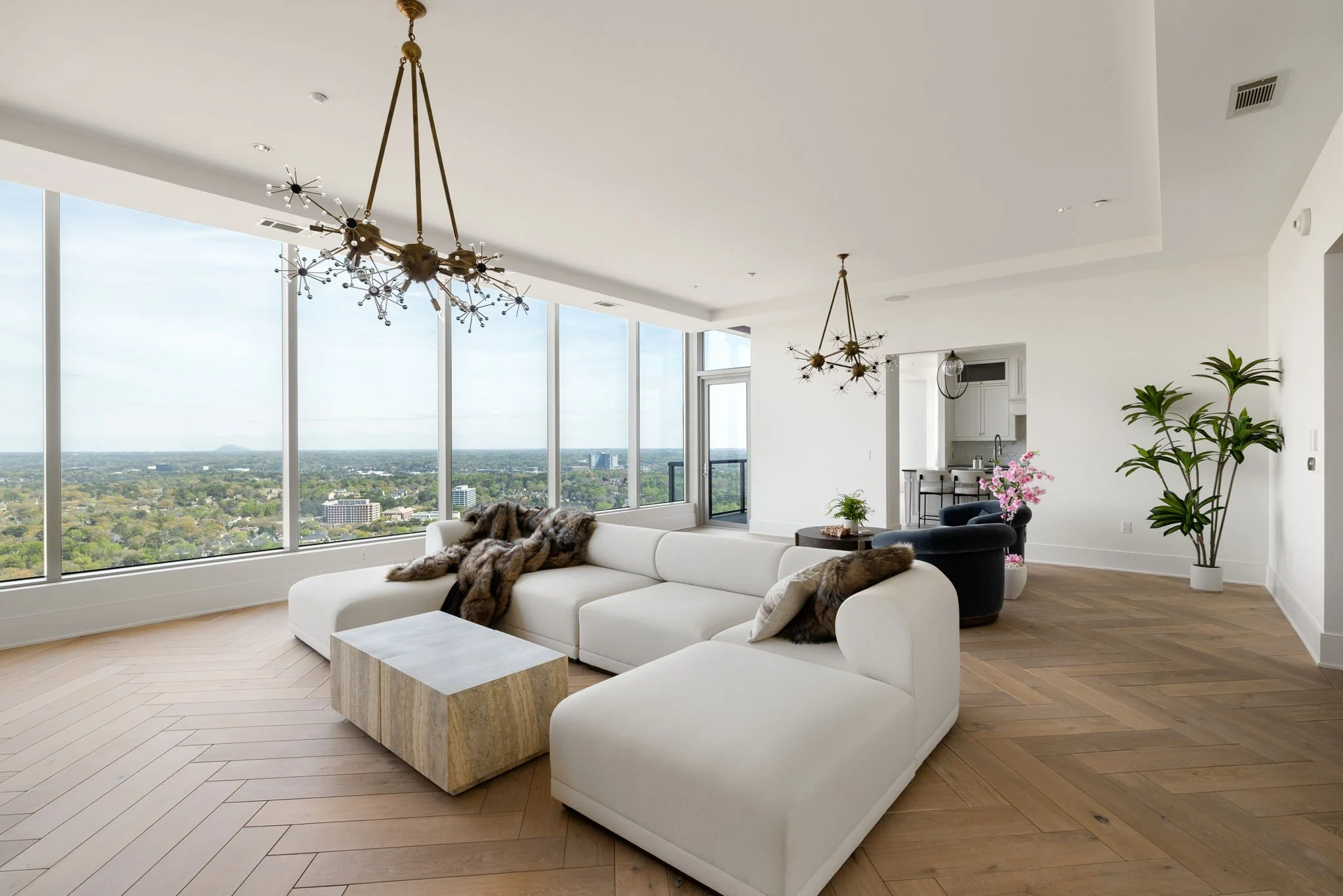 Modern living room with white sofa, fur throws, large windows, wooden flooring, black armchairs, pink flowers, and tall green plants, with a view of the city.
