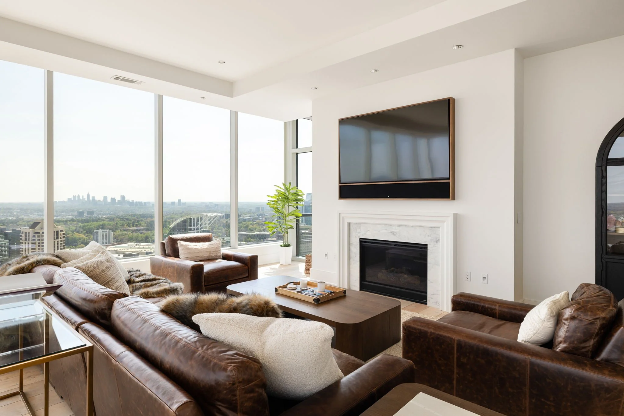 Modern living room with large windows, leather sofas, a fireplace, and a mounted TV, overlooking a city skyline.