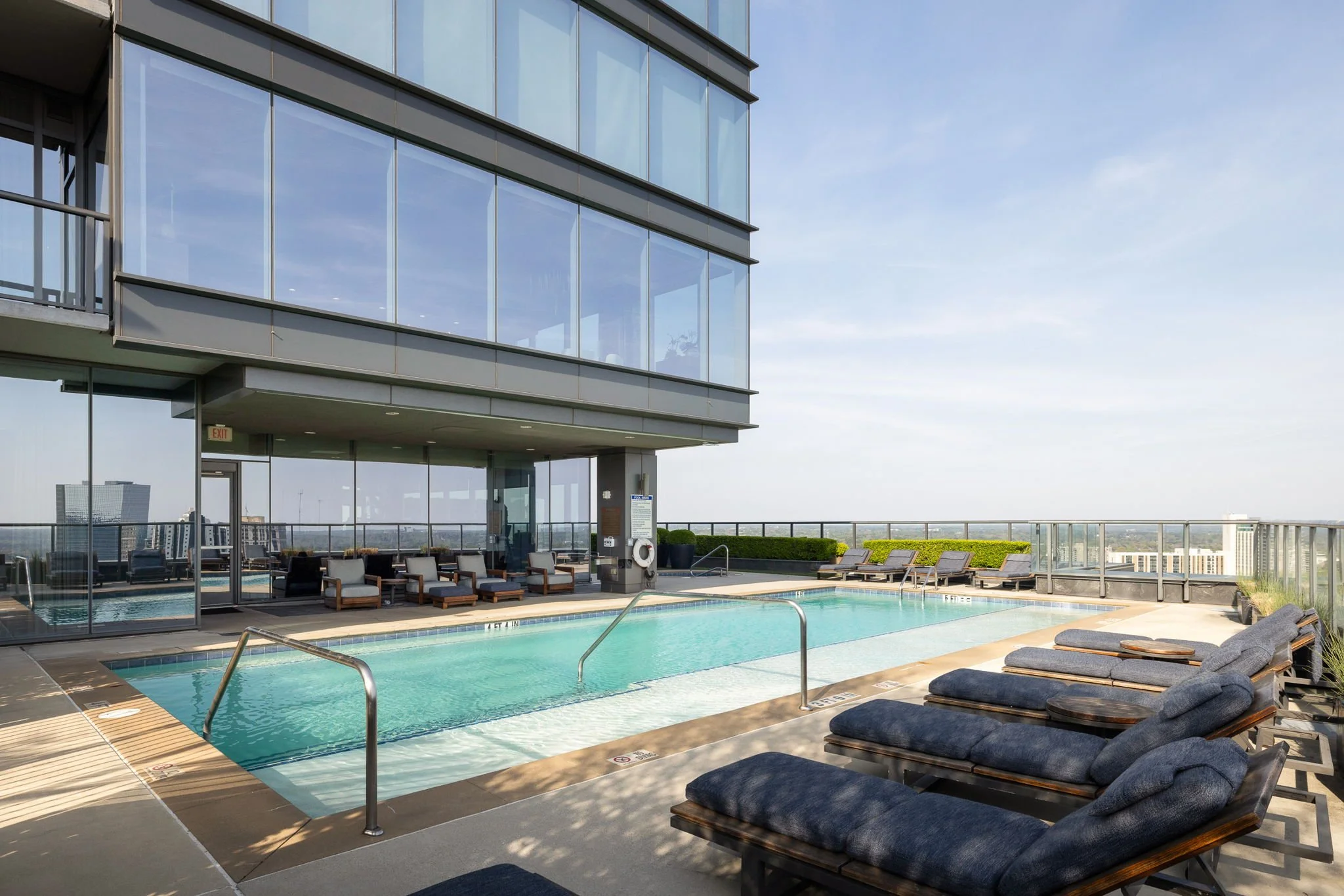 Rooftop swimming pool with lounge chairs, glass railings, and city view, adjacent to a modern glass building.