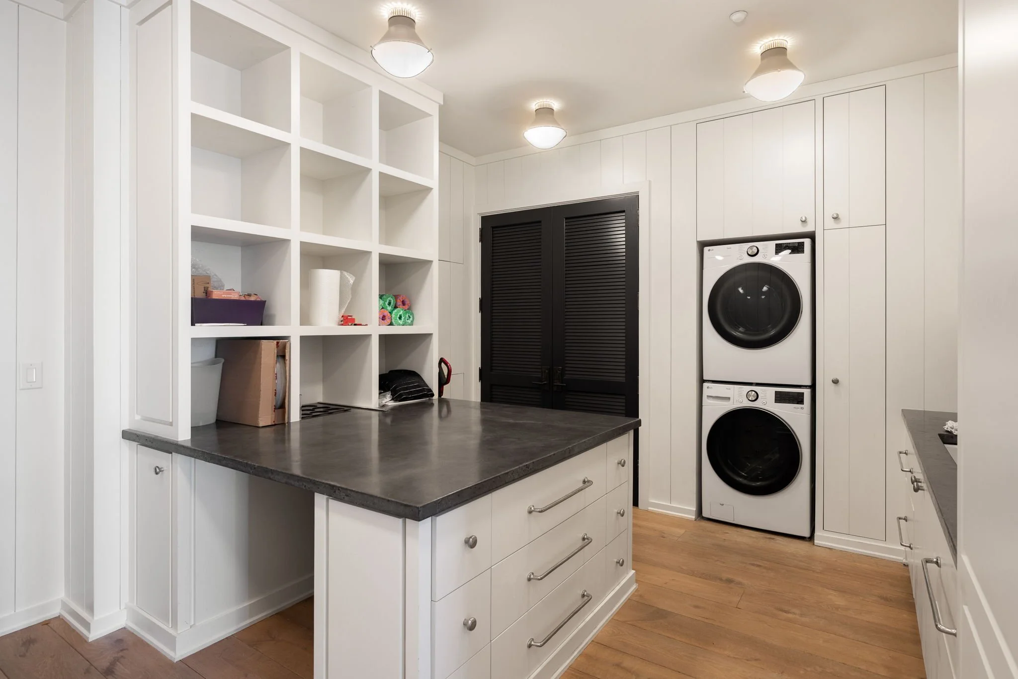 Laundry room with white cabinetry, a stacked washer and dryer, and open shelving with organized household items, hardwood flooring, and ceiling lights.