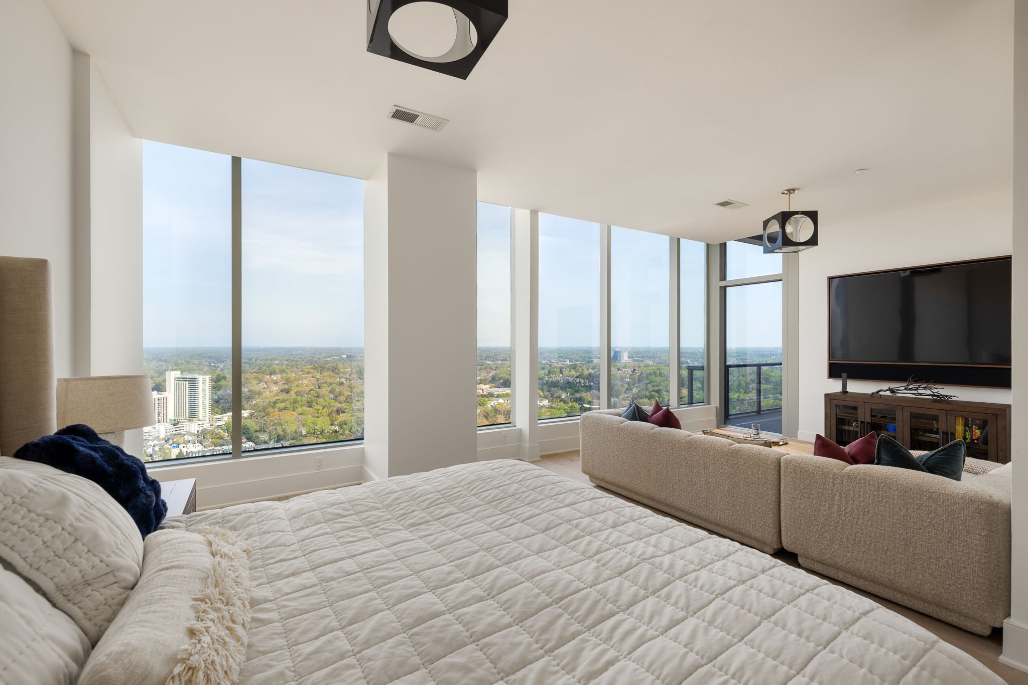 Modern bedroom with large windows providing a city view, a bed with beige bedding, a beige sofa with decorative pillows, and a flat-screen TV on the wall.