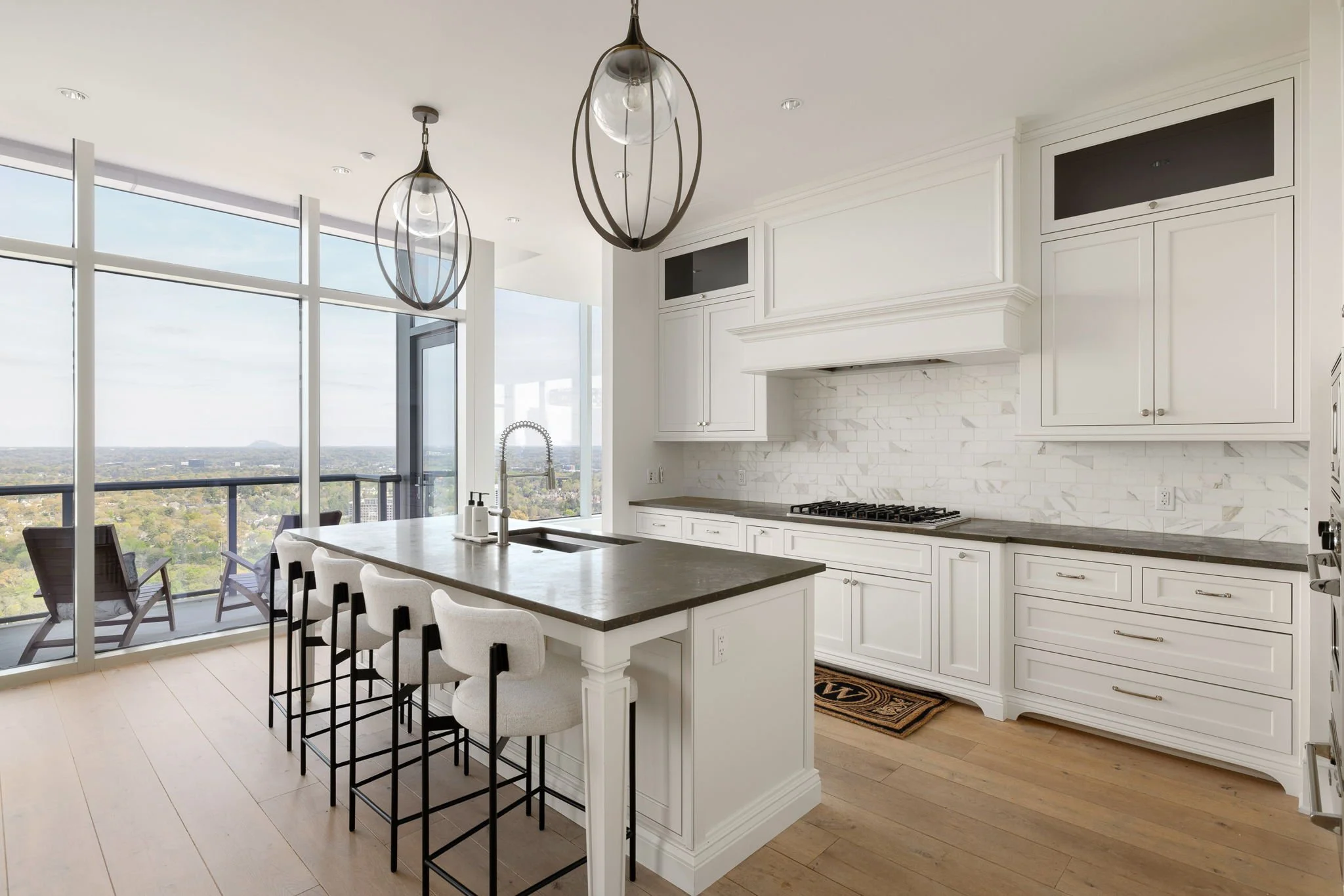 Modern kitchen with white cabinets, marble backsplash, dark countertop, island with seating, large windows showing city view, hanging pendant lights.