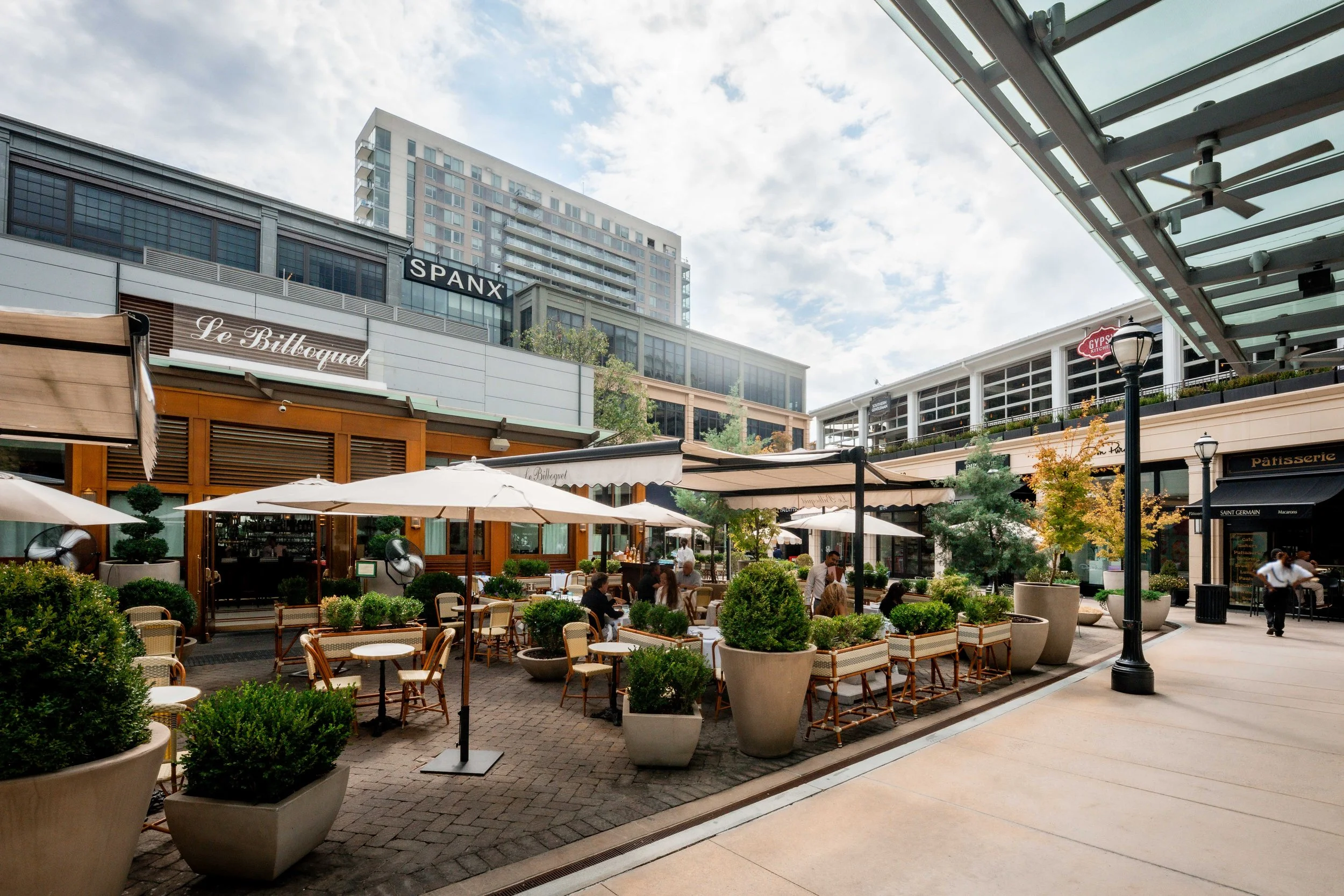 Outdoor shopping and dining area with tables, chairs, umbrellas, potted plants, and people, surrounded by modern buildings and storefronts under a partly cloudy sky.