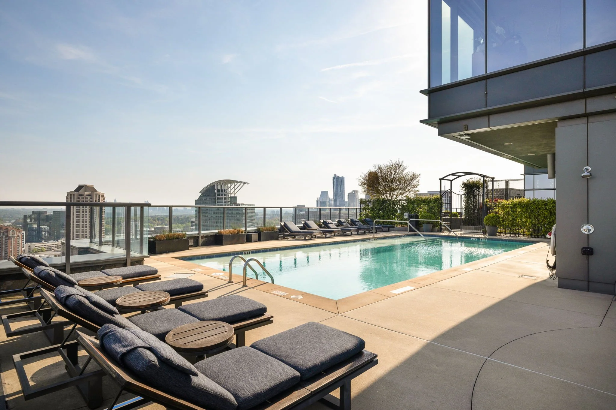 A rooftop swimming pool area with lounge chairs, potted plants, and city skyline views on a sunny day.