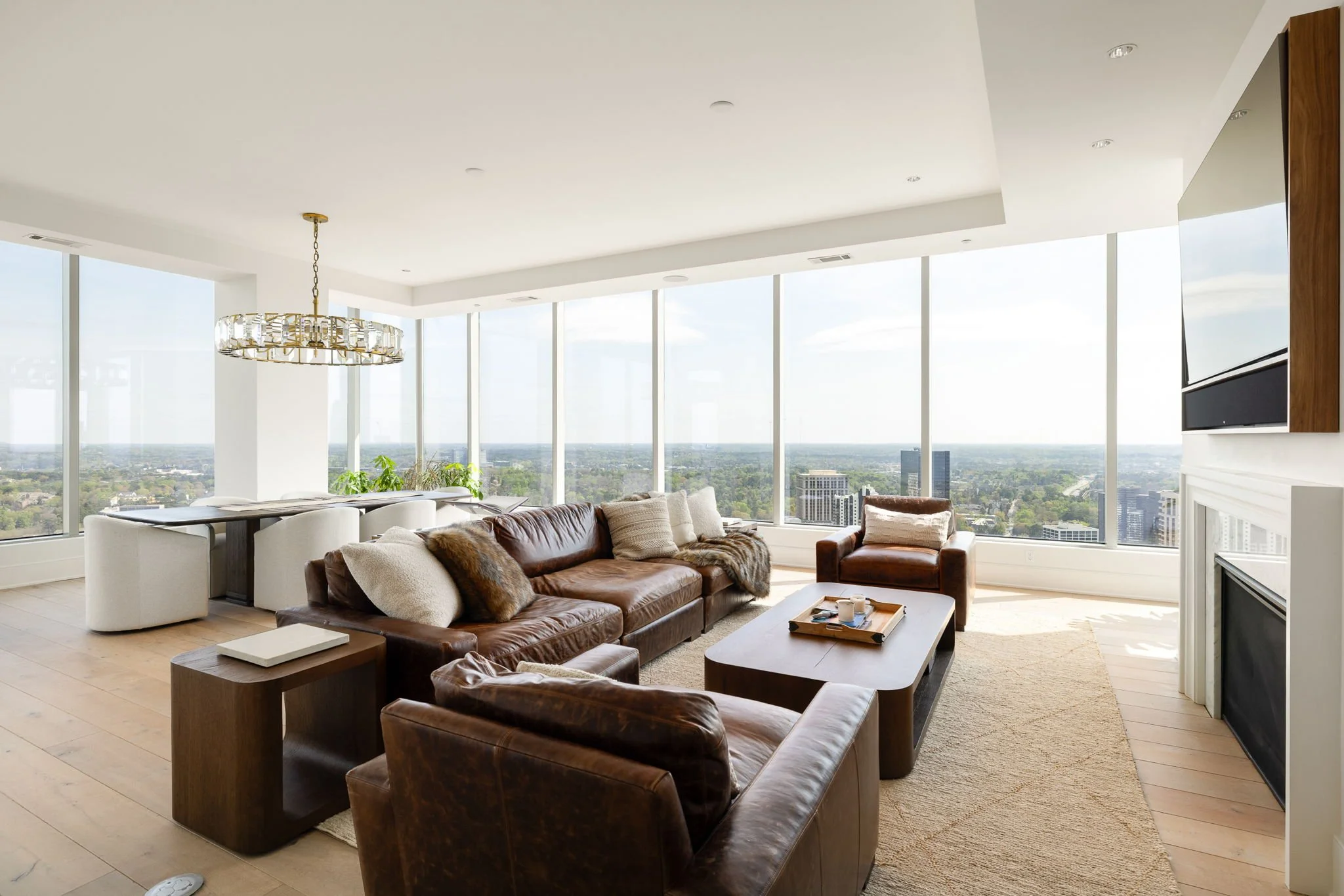 Modern living room with leather sofas, a wooden coffee table, large floor-to-ceiling windows with city views, a white fireplace, and an elegant chandelier.