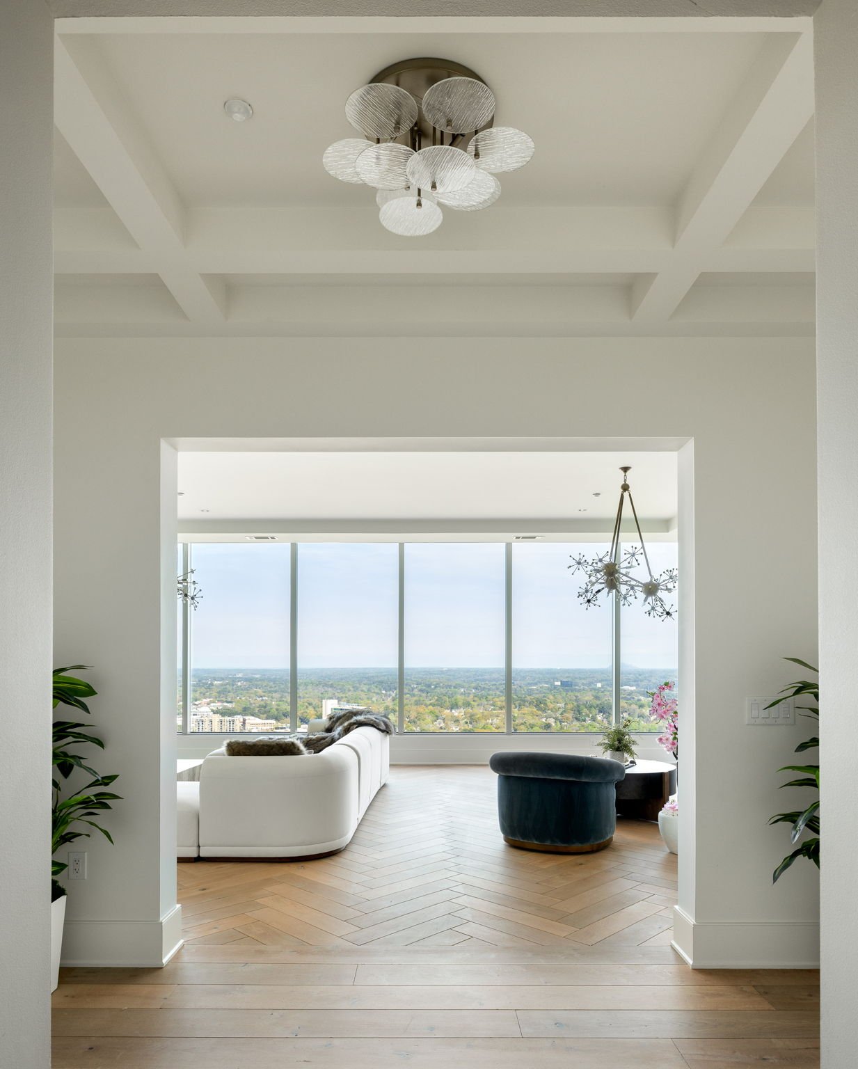 Bright living room with large floor-to-ceiling windows, white sofa, dark blue lounge chair, modern chandelier, and greenery.