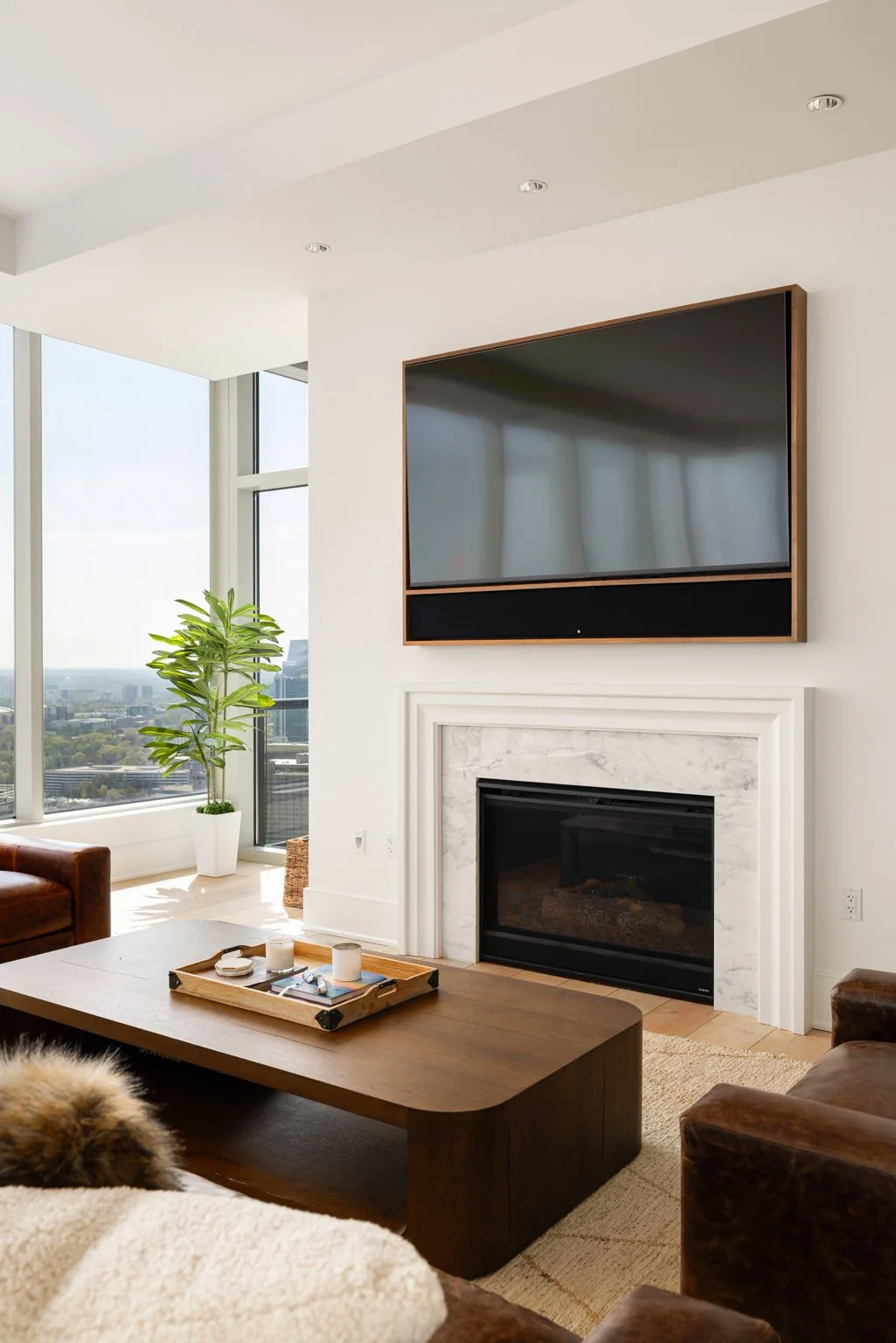 Living room with a large flat-screen TV above a fireplace, a wooden coffee table with candles, a potted plant by the window, and brown leather chairs.