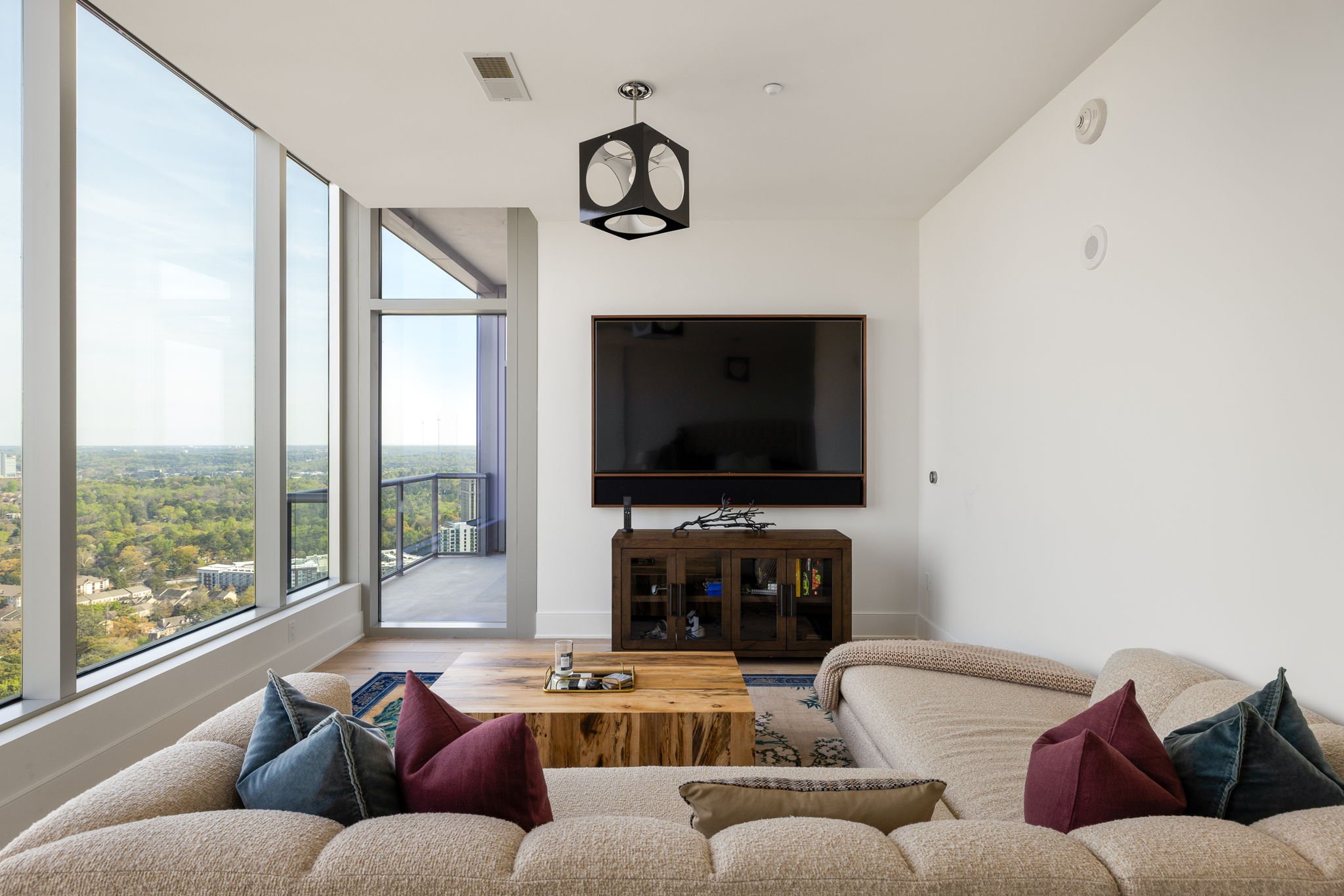 Living room with large windows showing city view, beige sectional sofa with blue and red pillows, wooden coffee table, TV mounted on white wall with a dark wooden cabinet underneath, small balcony door, modern ceiling light fixture.
