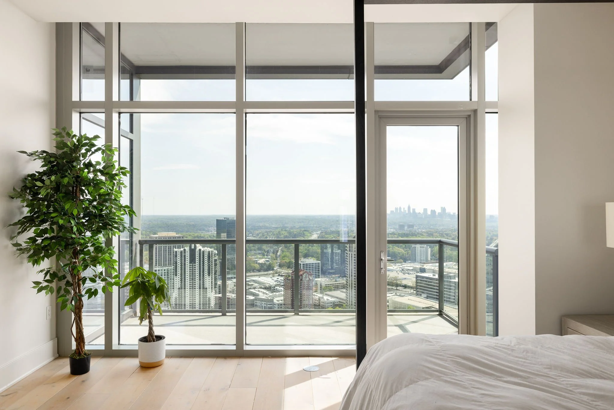 Interior of a modern high-rise apartment bedroom with large floor-to-ceiling windows, overlooking a city skyline with tall buildings and green landscape, with a bed in the foreground and two large potted plants near the window.