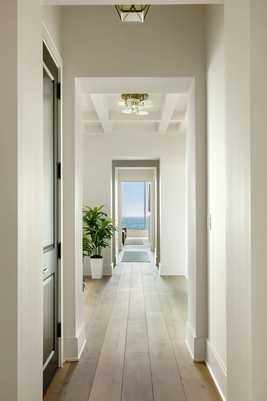 Bright hallway with white walls, light wooden flooring, a plant in a white pot, and a large window with a view of the sky and horizon at the end.