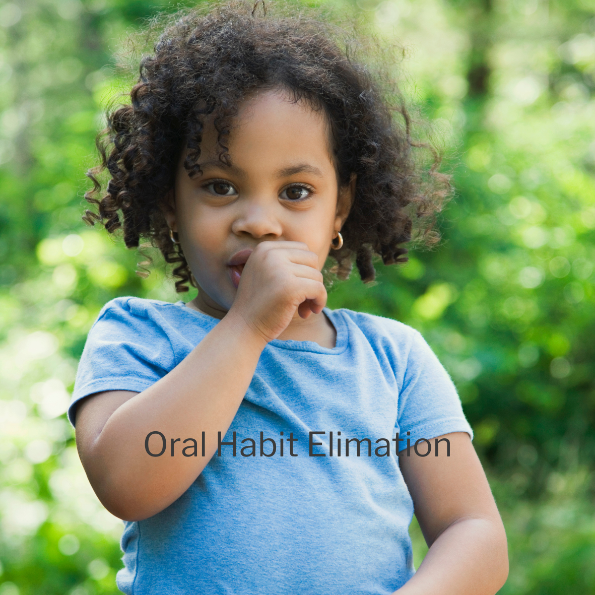 Young girl with curly hair wearing a blue shirt, standing outdoors with green trees in the background, holding her hand near her mouth, illustrating oral habit elimination.