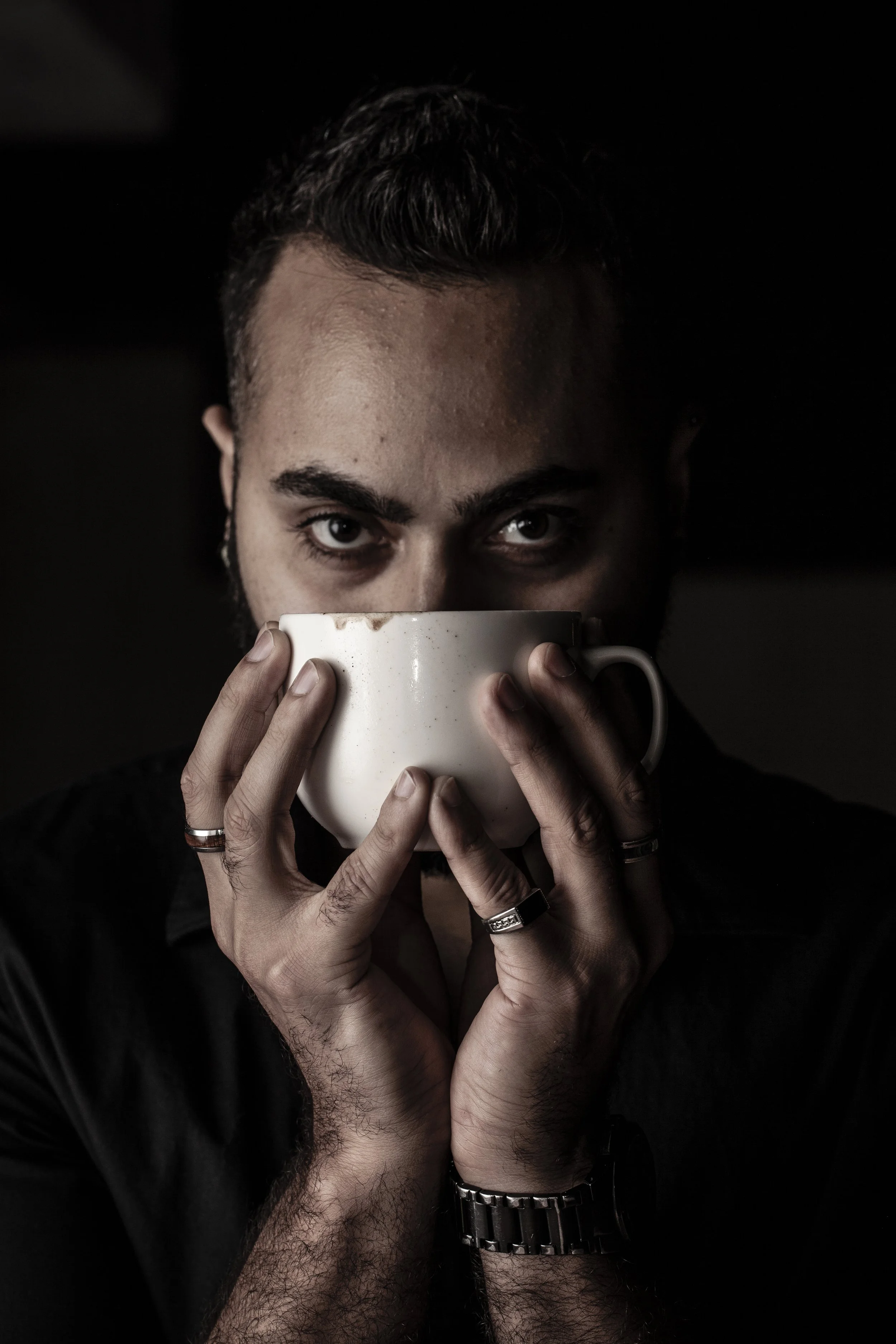 A man with dark hair and facial hair holding a large white mug, partially covering his face, with intense eyes staring forward.