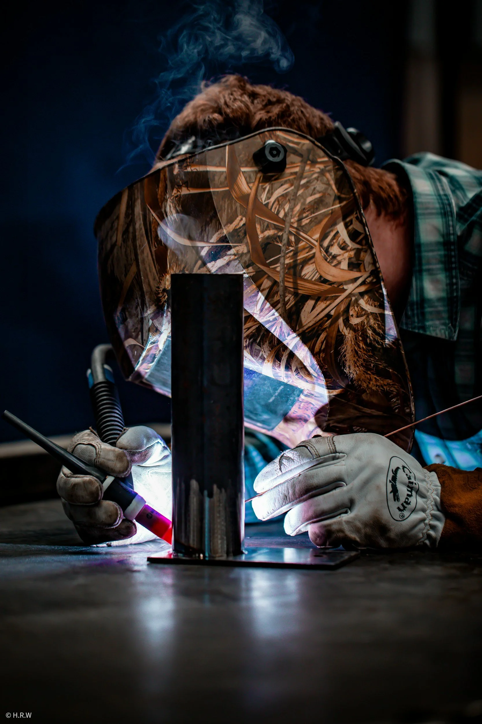 A welder wearing a protective helmet, gloves, and plaid shirt welding a metal structure with sparks and light.
