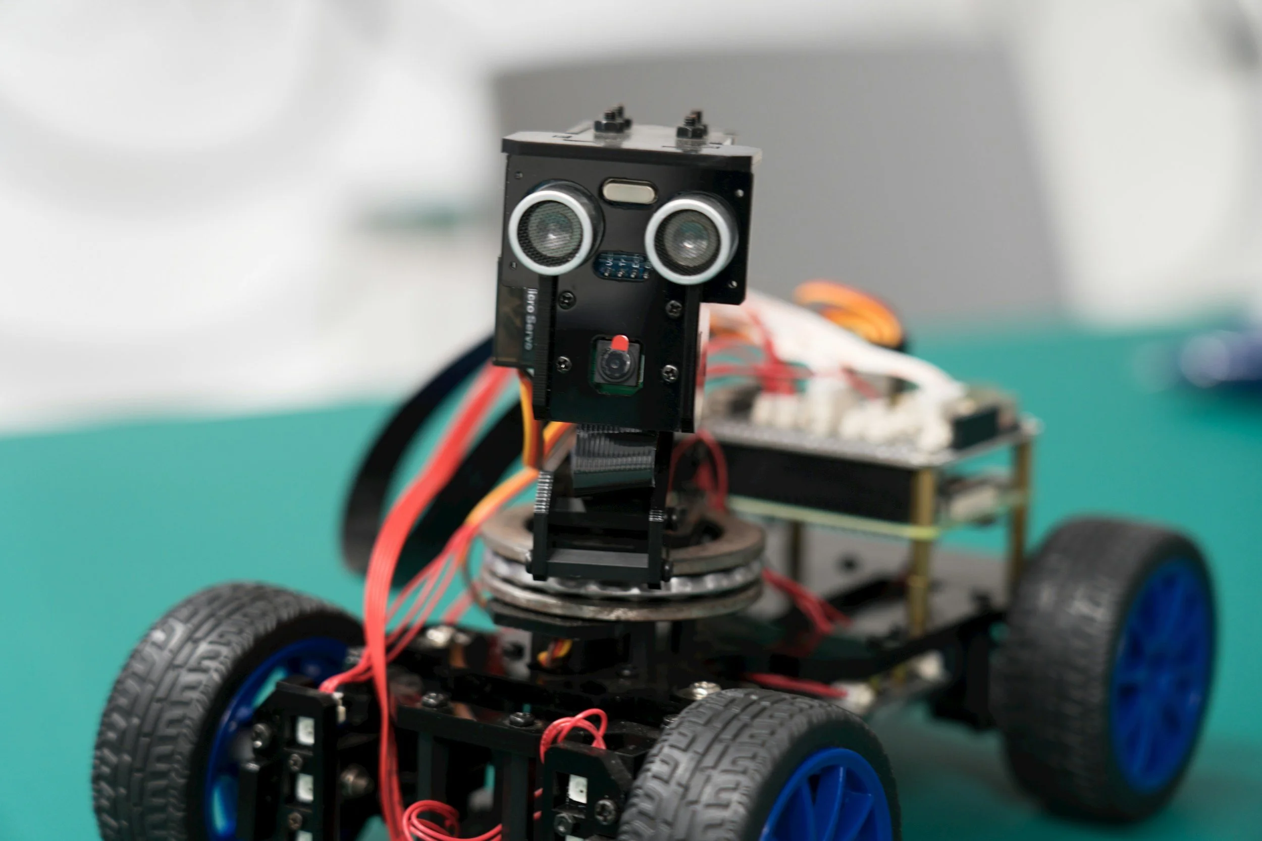 A small robot with four wheels, wires, and electronic components, featuring a camera and speaker grill on its face.