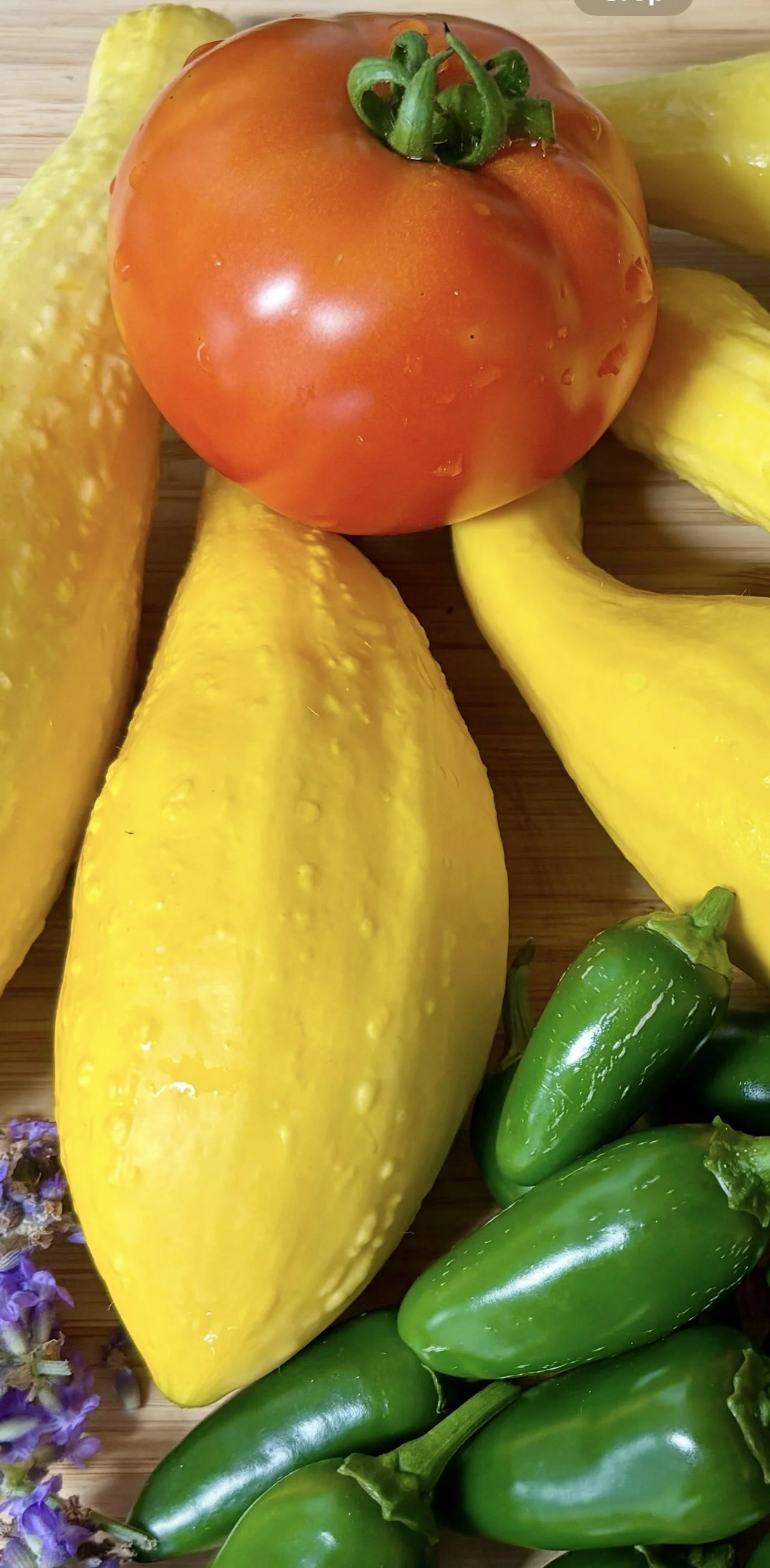 A red tomato, yellow summer squash, small green jalapeño peppers, and purple flowers on a wooden surface.
