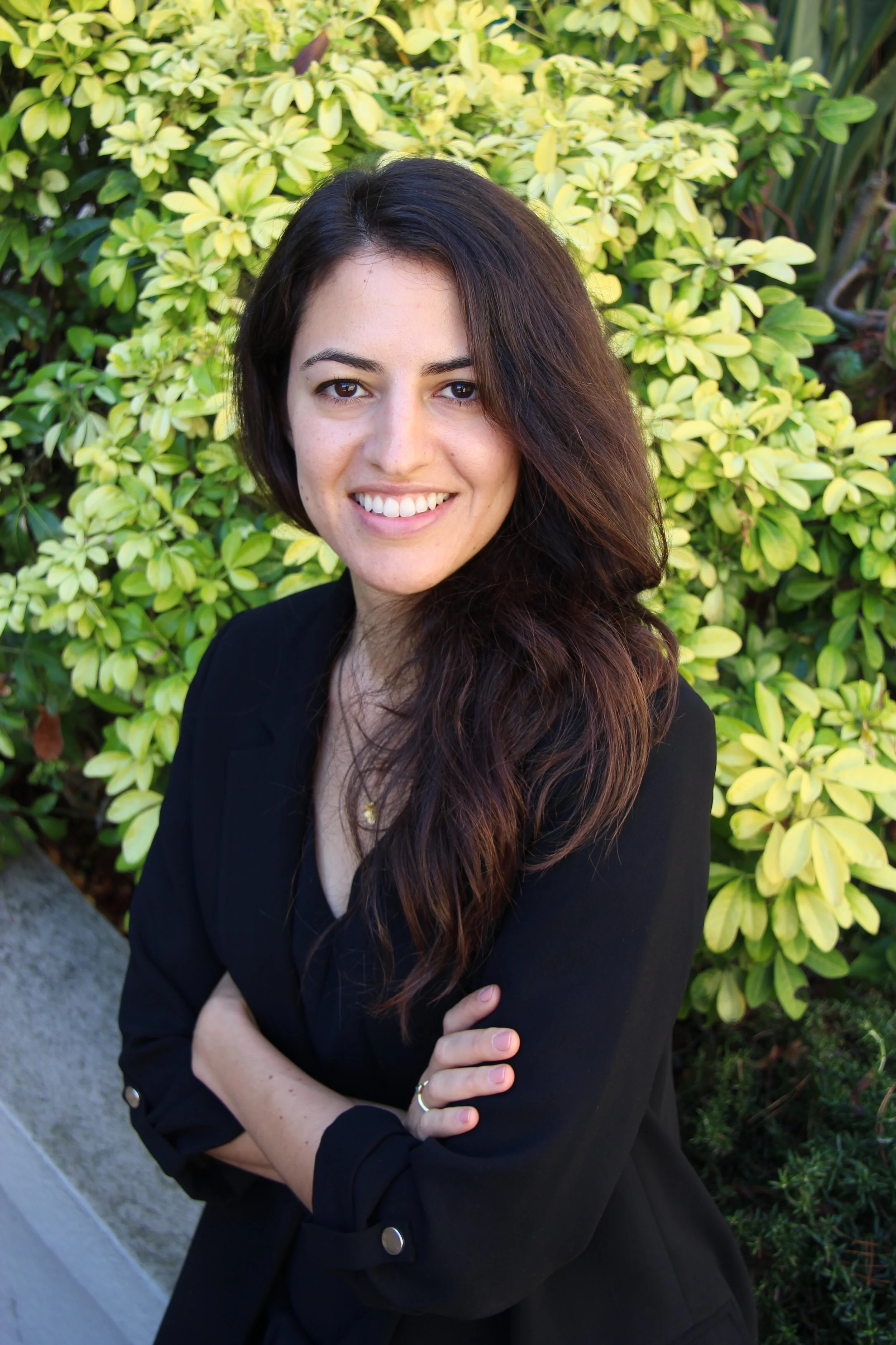 A woman with long wavy dark hair, wearing a black blazer, standing outdoors in front of green leafy bushes, smiling and crossing her arms.