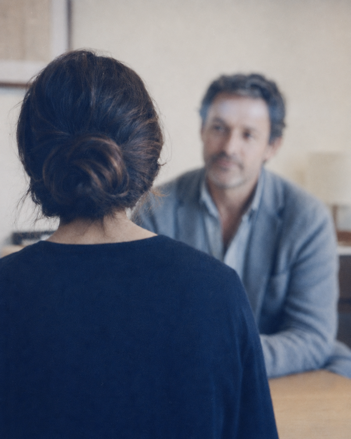 A woman with her hair in a low bun sits across from a softly out-of-focus man during a professional conversation.