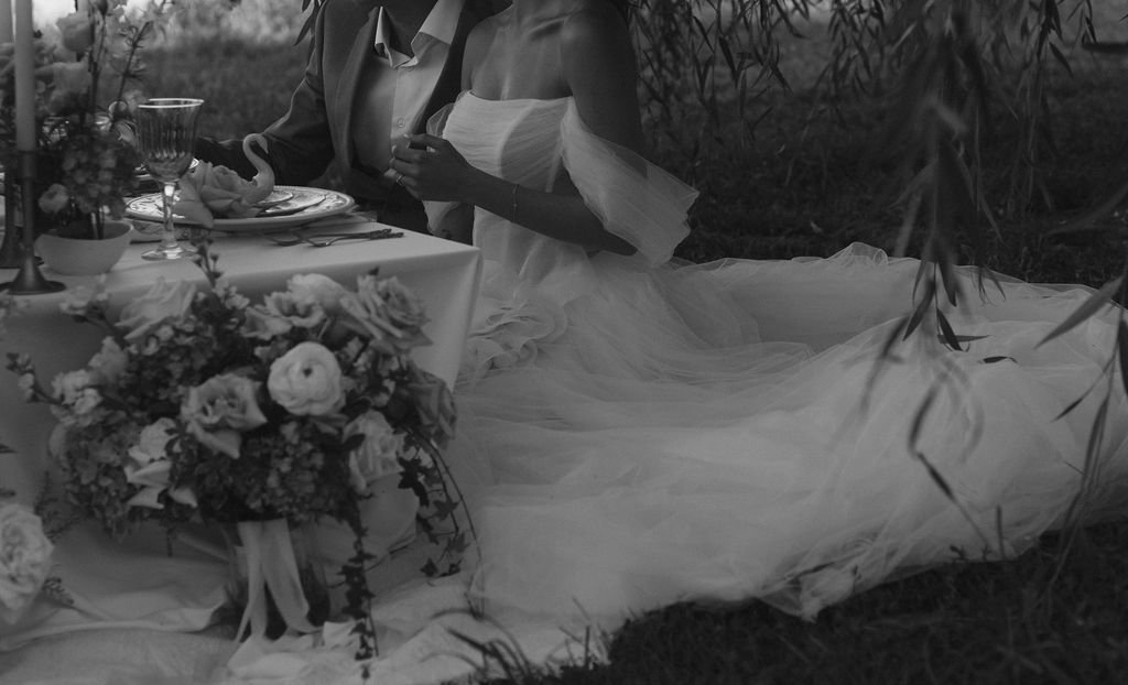 A bride and groom sitting on the grass for a romantic outdoor wedding celebration, with a table of food and floral arrangements in the foreground.