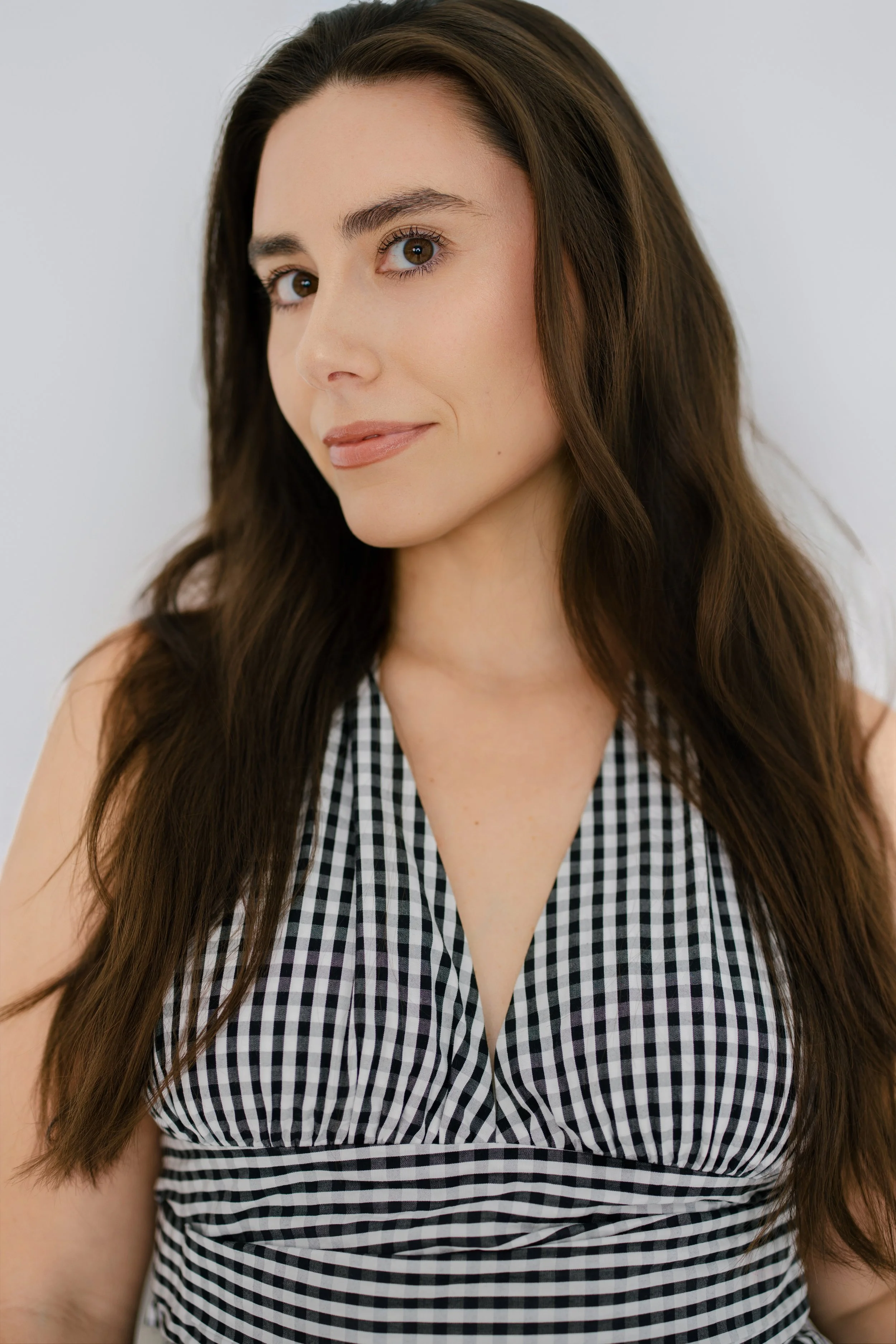 Close-up of a young woman with long brown hair and light skin, wearing a black and white gingham sleeveless top, against a plain white background.